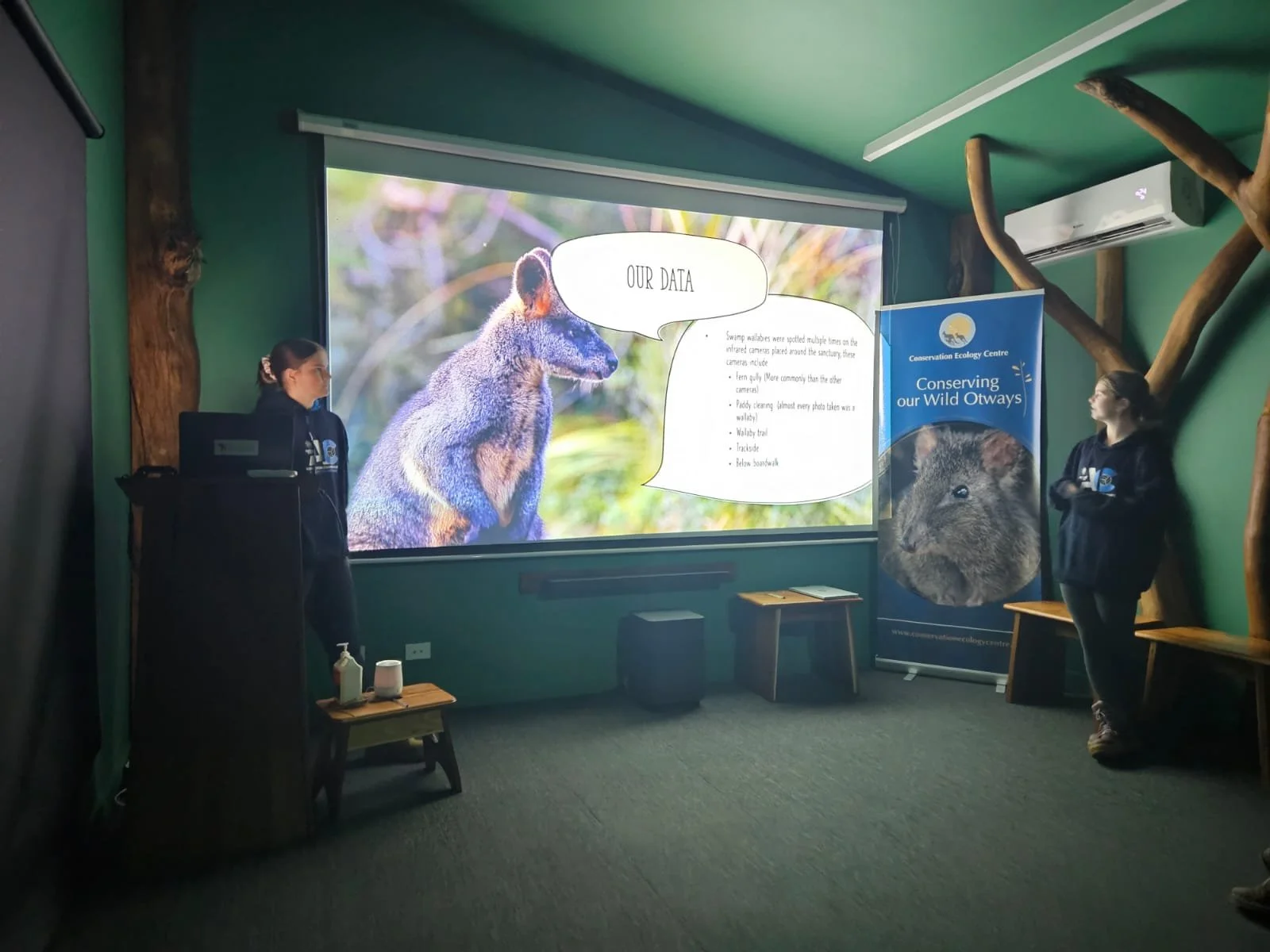 Two presenters in a room with a large screen displaying wildlife information about swamp wallabies, part of a presentation at a conservation center. A banner beside them reads "Conserving our Wild Otways." The space is decorated with wooden elements