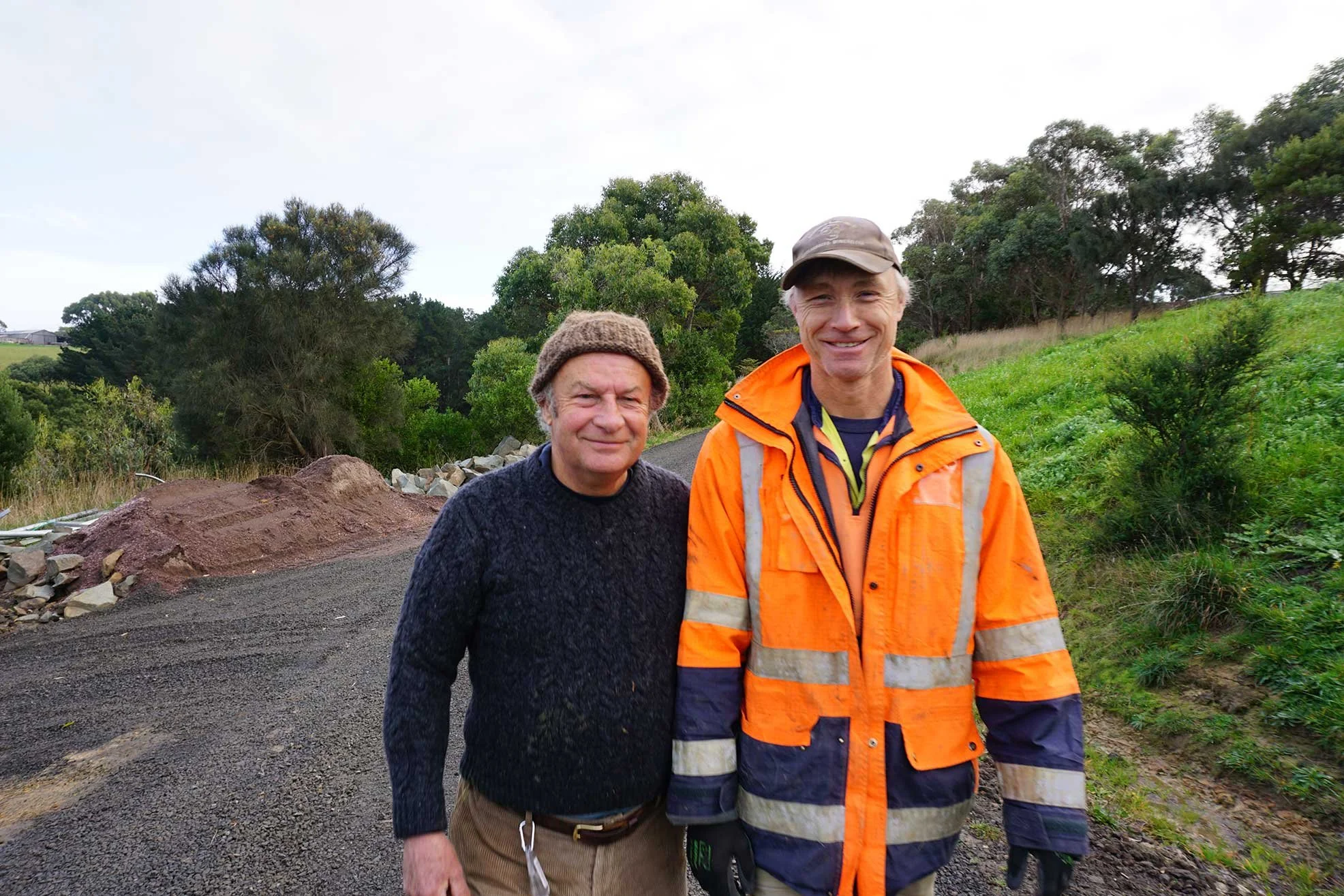 Two men standing on a gravel path in a rural outdoor setting, one wearing a hat and sweater, the other in an orange safety jacket.