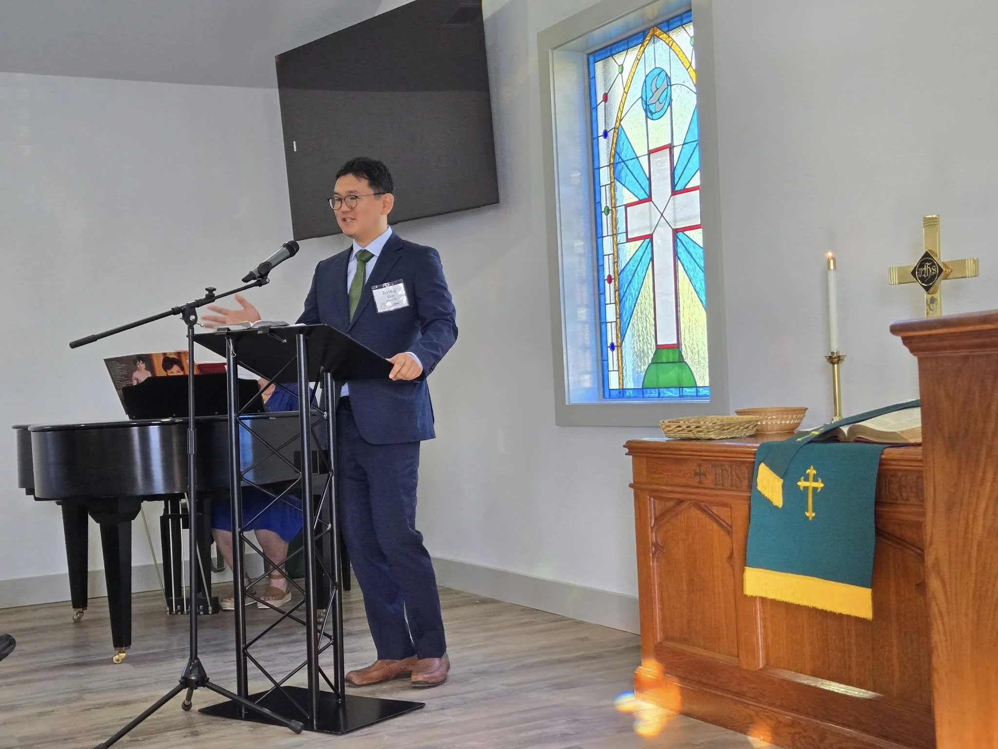 A man in a blue suit and green tie speaking at a podium in a church, with a woman playing a piano behind him. There is stained glass with a cross and dove design, candles, and religious items on a wooden altar.