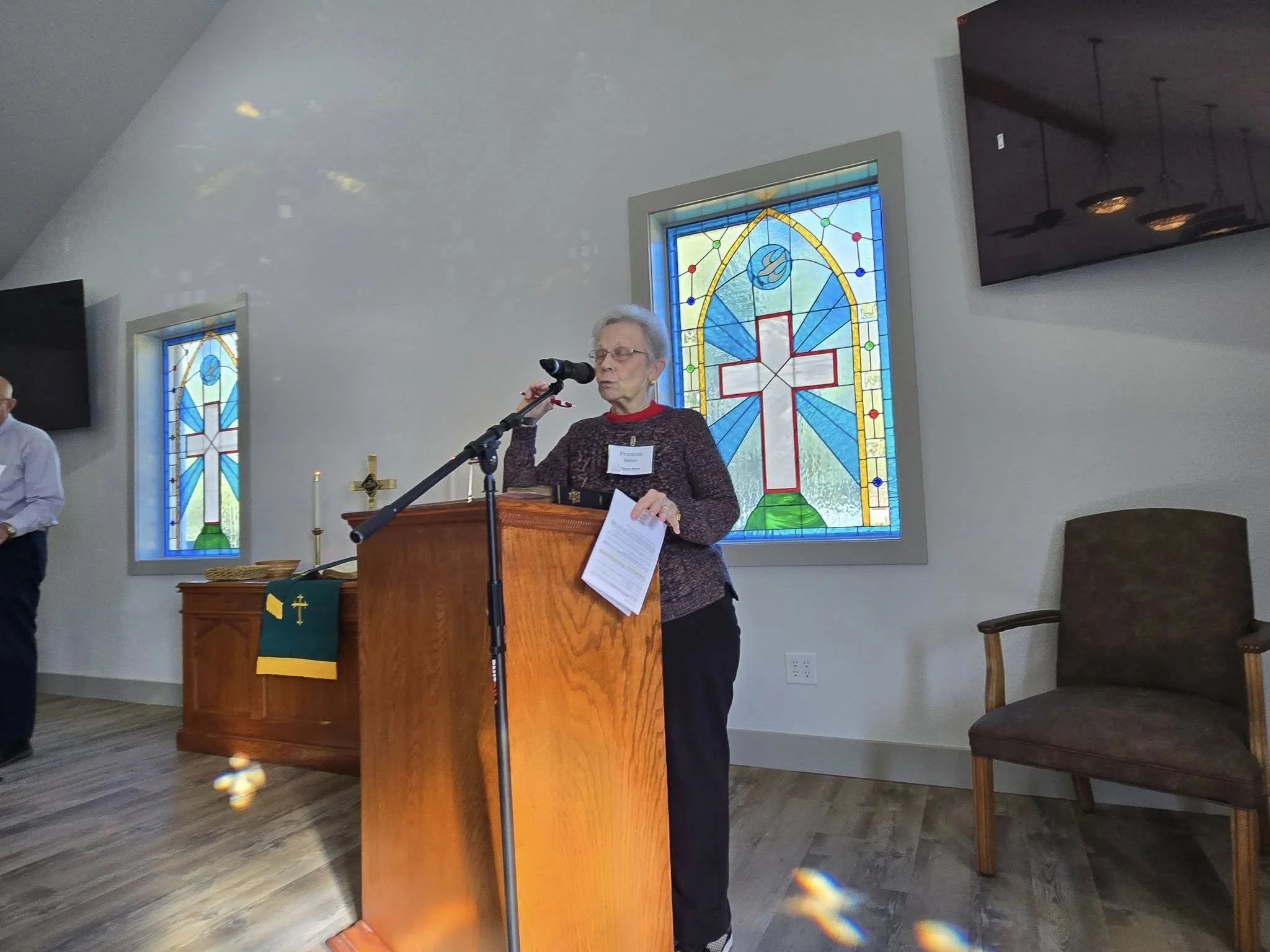 An elderly woman with glasses speaking at a podium inside a church, with stained glass windows in the background depicting a cross and abstract colorful designs.