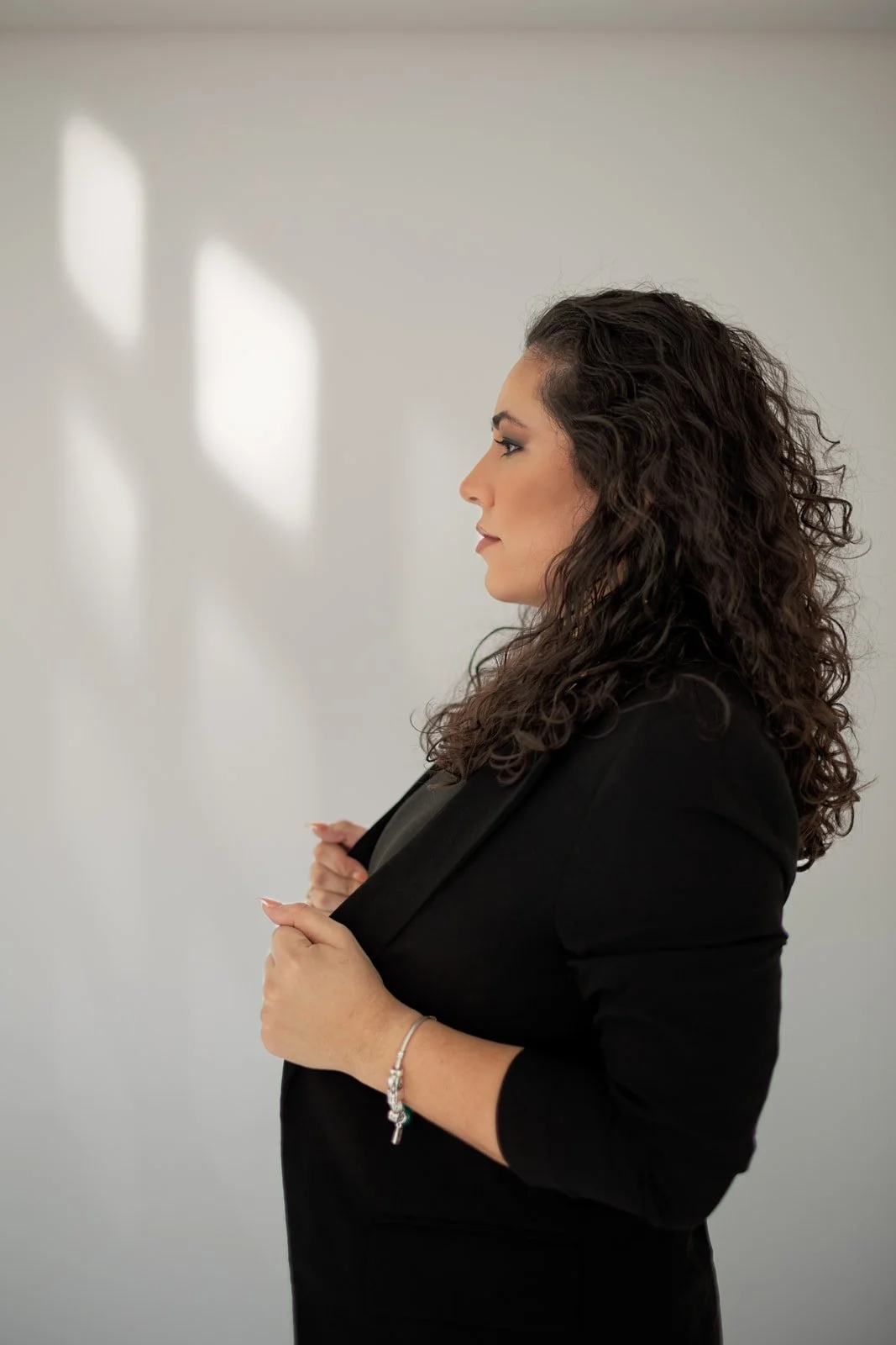Profile of a woman with curly dark hair wearing a black blazer, adjusting her blazer with her hands, standing against a light background with soft political shadows. Orlando-florida-wedding-videographer-portrait