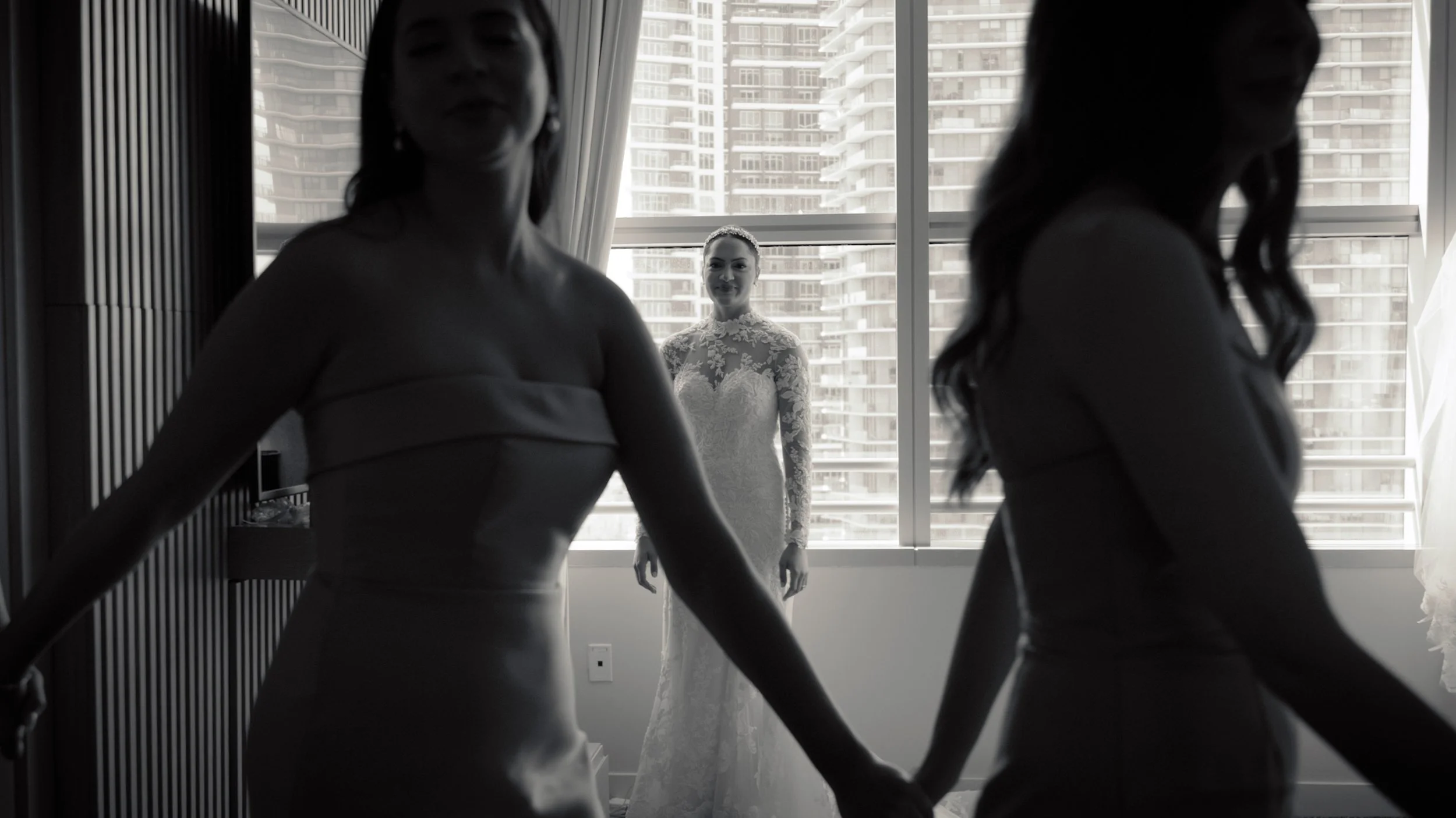 Two women holding hands in silhouette in the foreground, with a bride looking out the window behind them, in a room with a cityscape view. Miami-florida-wedding-videographer