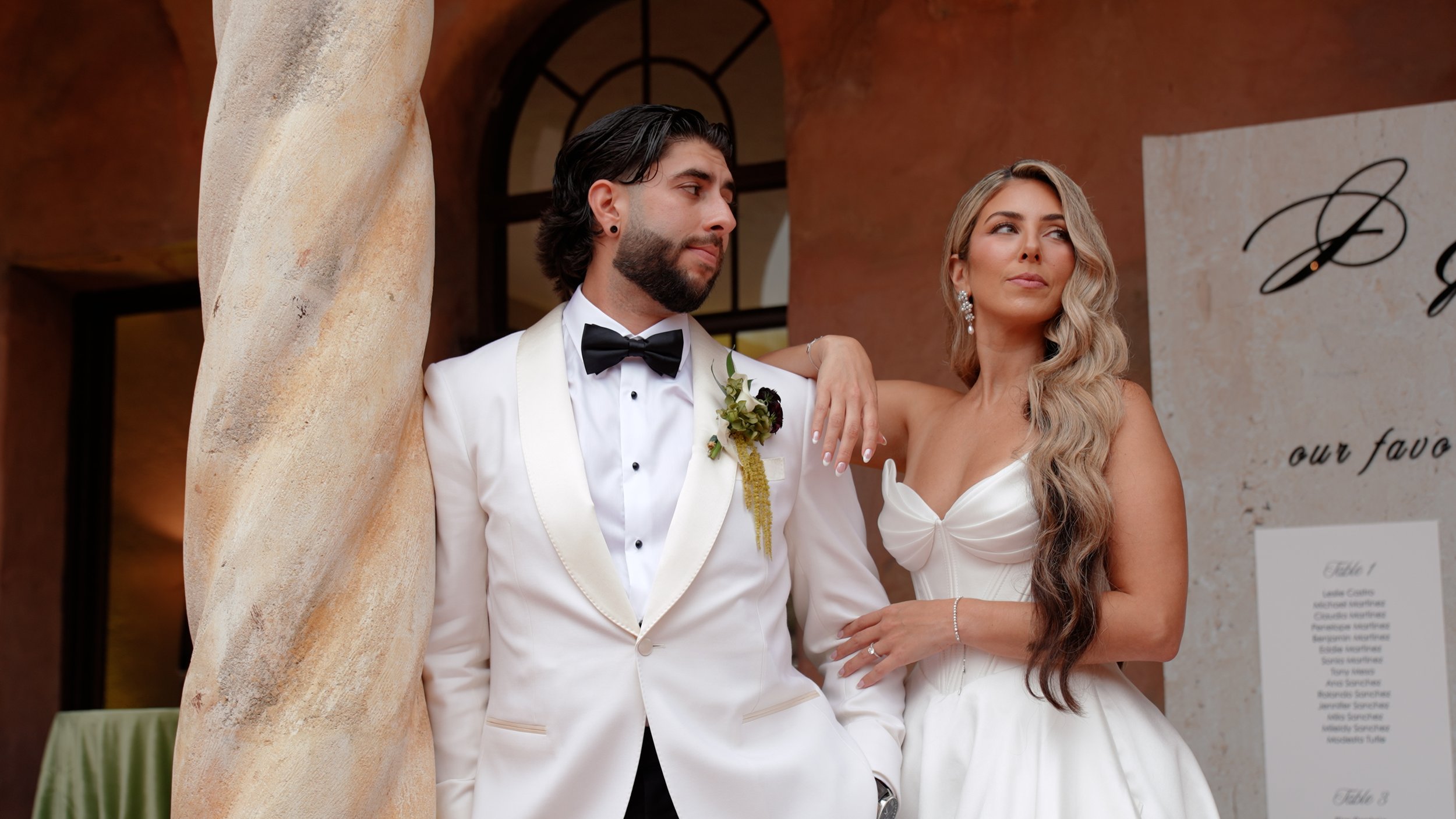 A groom and bride posing at their wedding, standing near a stone column in an elegant venue. The groom is wearing a white tuxedo with a black bow tie and boutonniere, and the bride is in a white strapless gown with long wavy hair, earrings, and a bracelet.