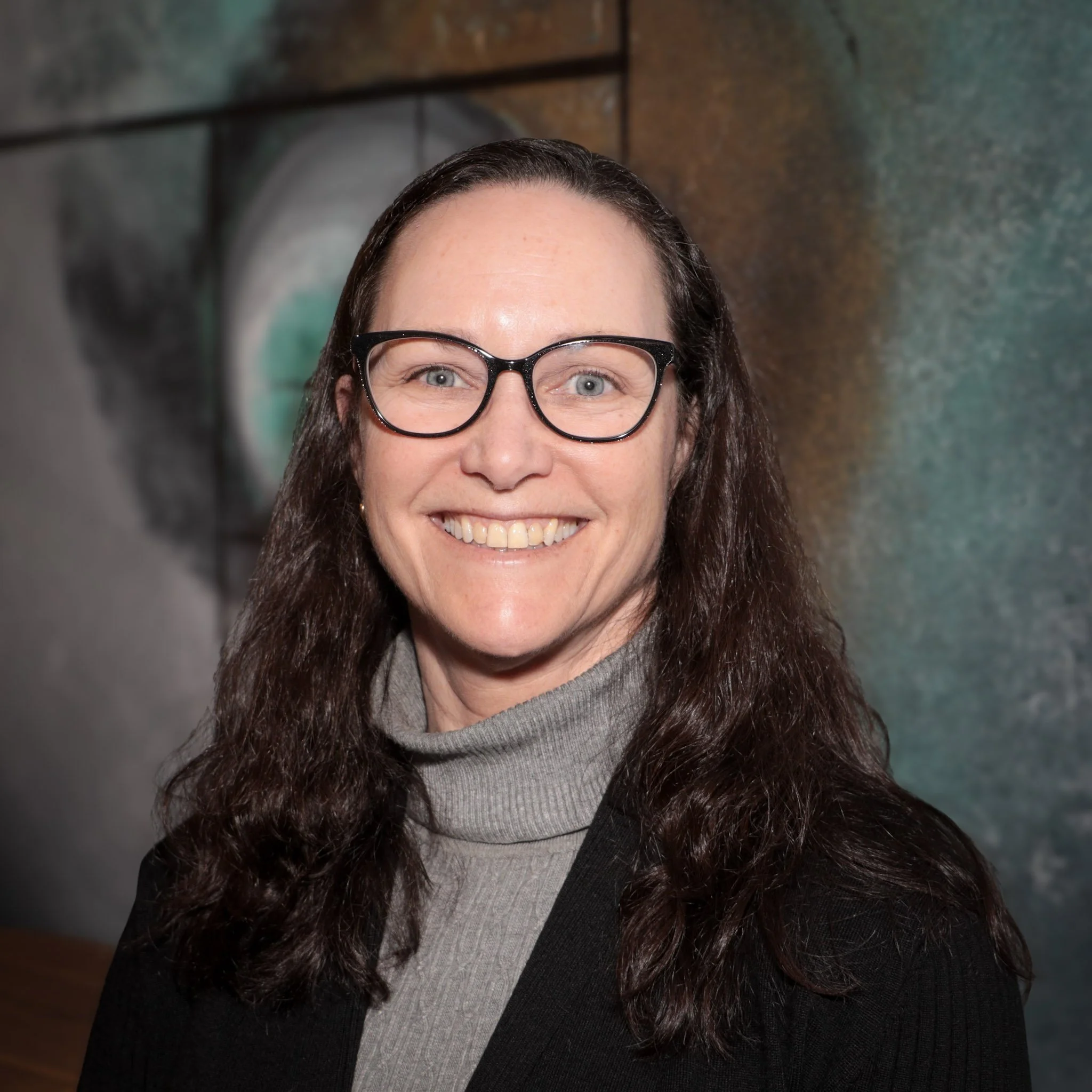 Smiling person with curly hair wearing a dark jacket and blue shirt against a white background.