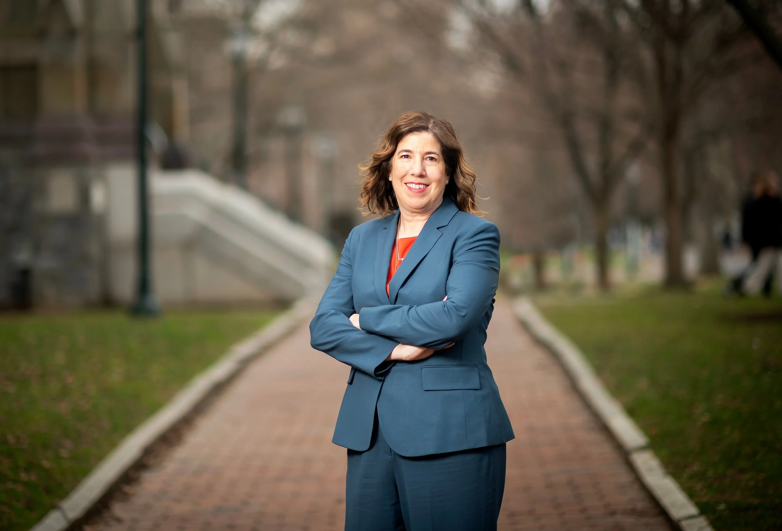 Smiling older person with gray hair wearing red glasses and a patterned sweater, against a gray background.