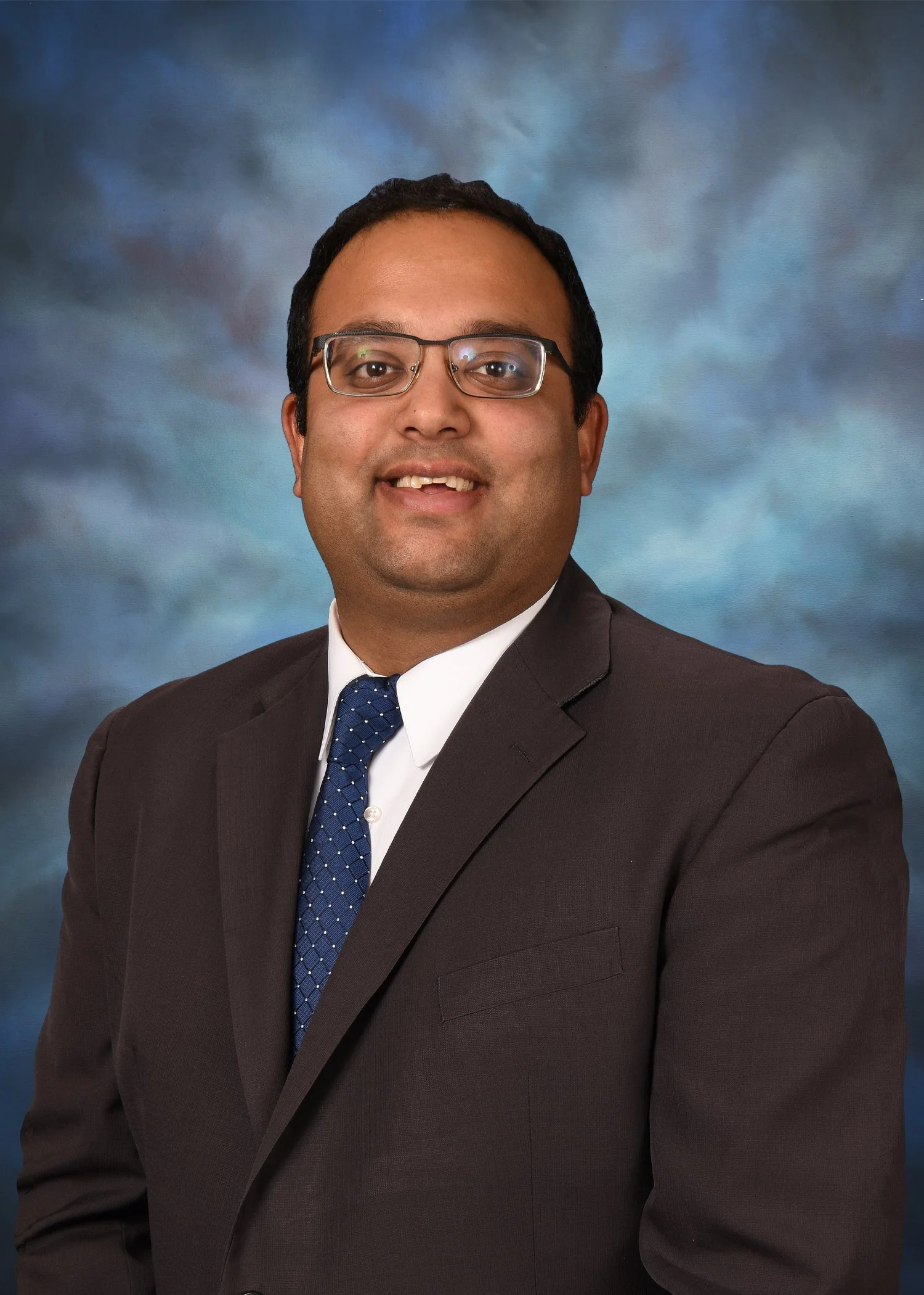 Person smiling wearing glasses and a gray sweater, standing in front of a colorful map background.