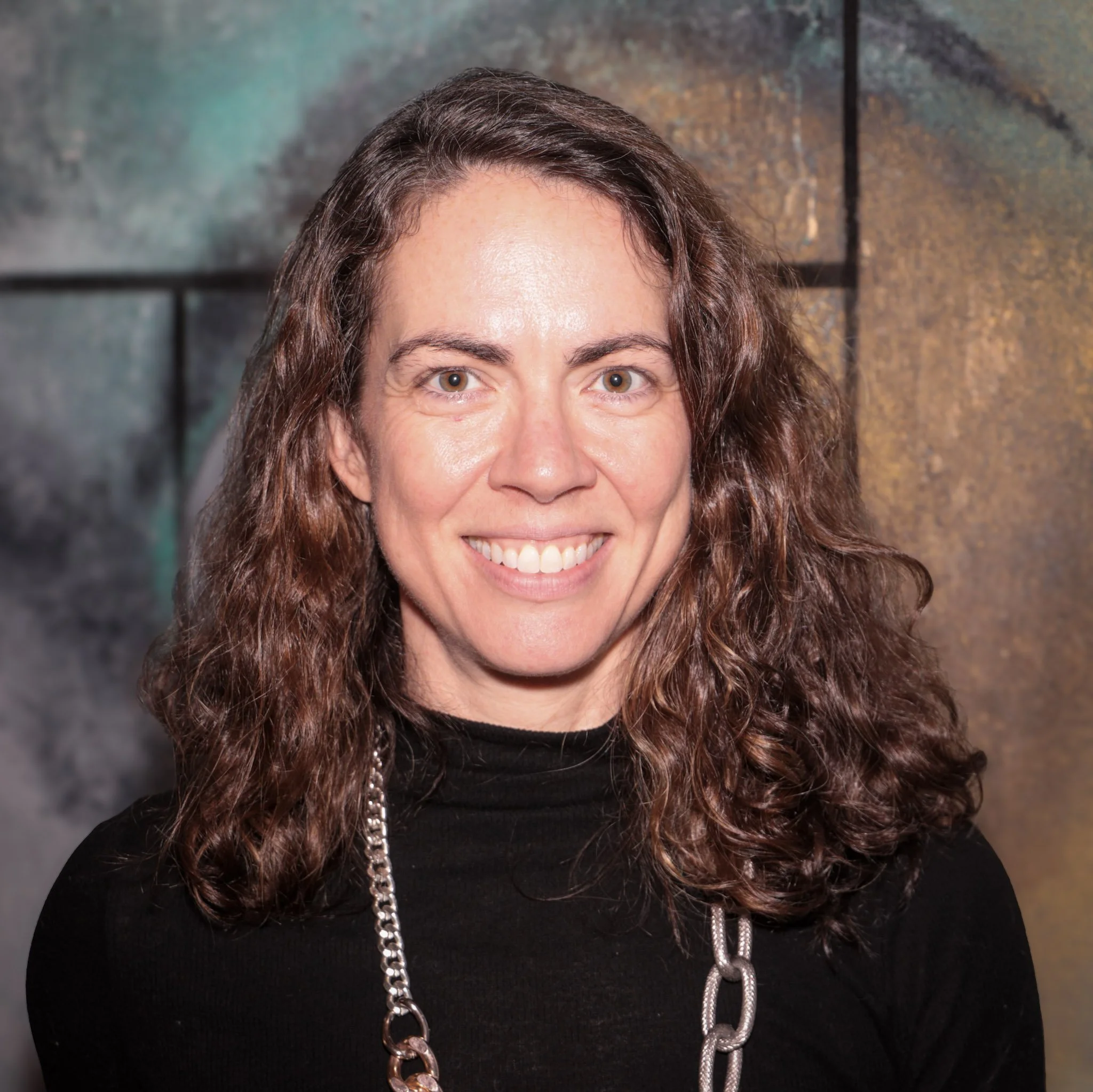 Smiling person with long dark hair, wearing a white shirt and necklace, on a white background.