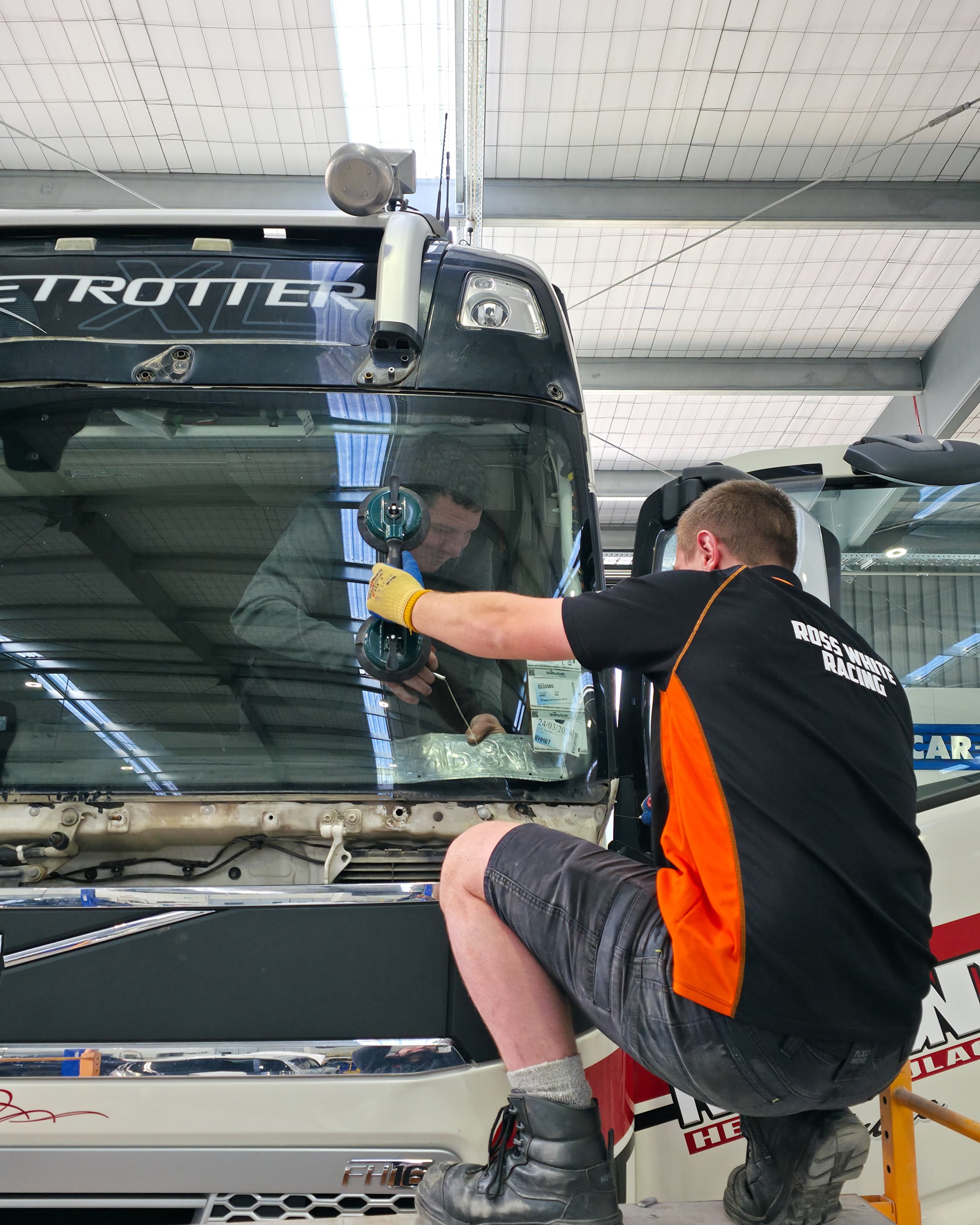 A mechanic working on a commercial truck inside a large industrial garage, with another person inside the truck's cab.