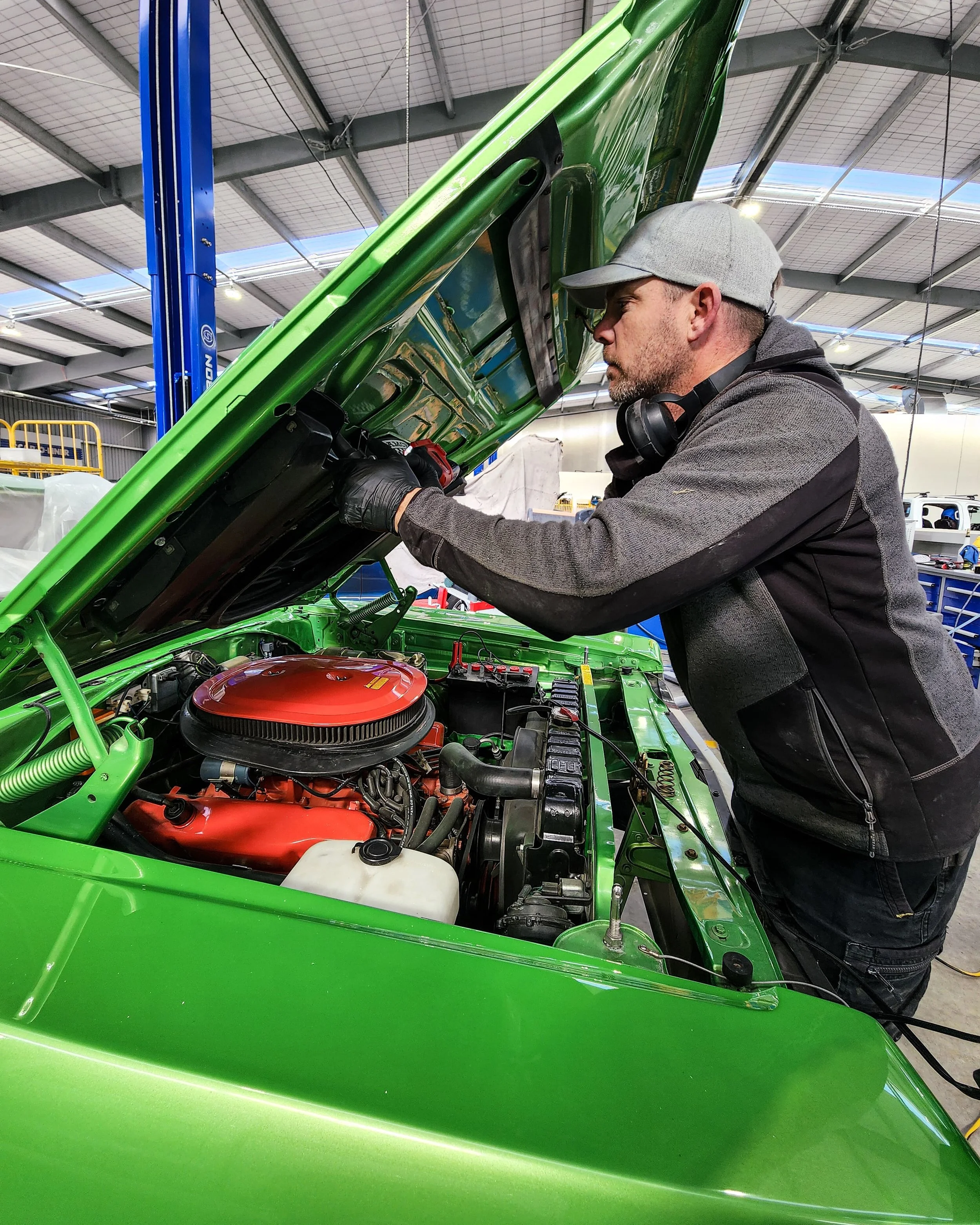 A man wearing a gray cap, gray and black jacket, and headphones around his neck is working on the engine of a bright green vintage car inside a garage or workshop. The hood of the car is open, revealing a red air filter and other mechanical component