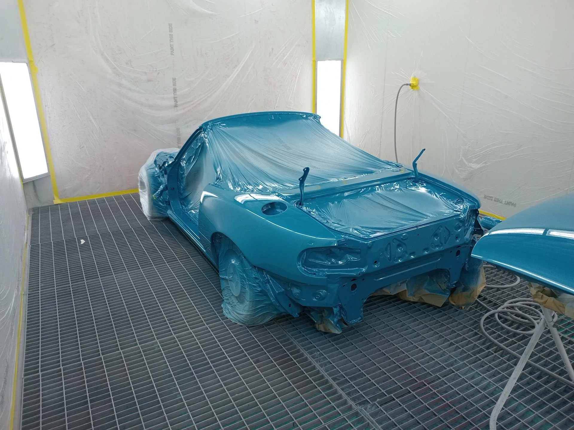 A car inside a paint booth, covered with blue protective plastic, being painted or coated.