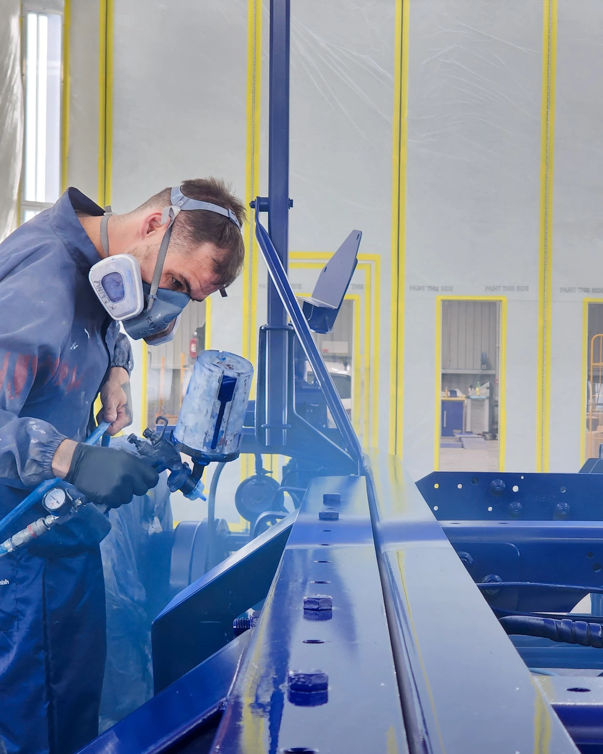 A worker wearing protective gear including a mask and gloves uses a spray gun on a metal structure in a workshop with yellow and blue markings.