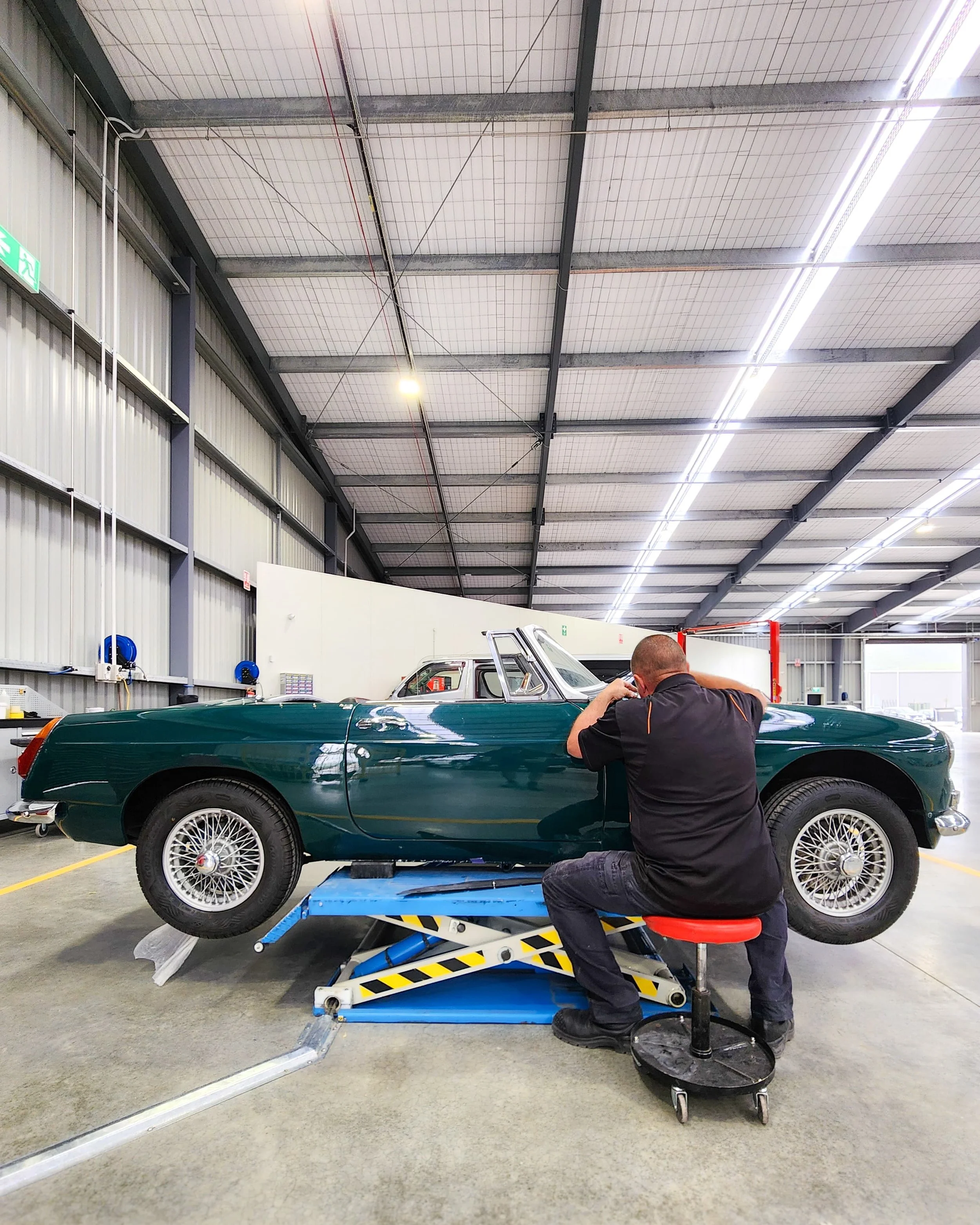 A man working on a vintage green car inside a spacious industrial workshop.