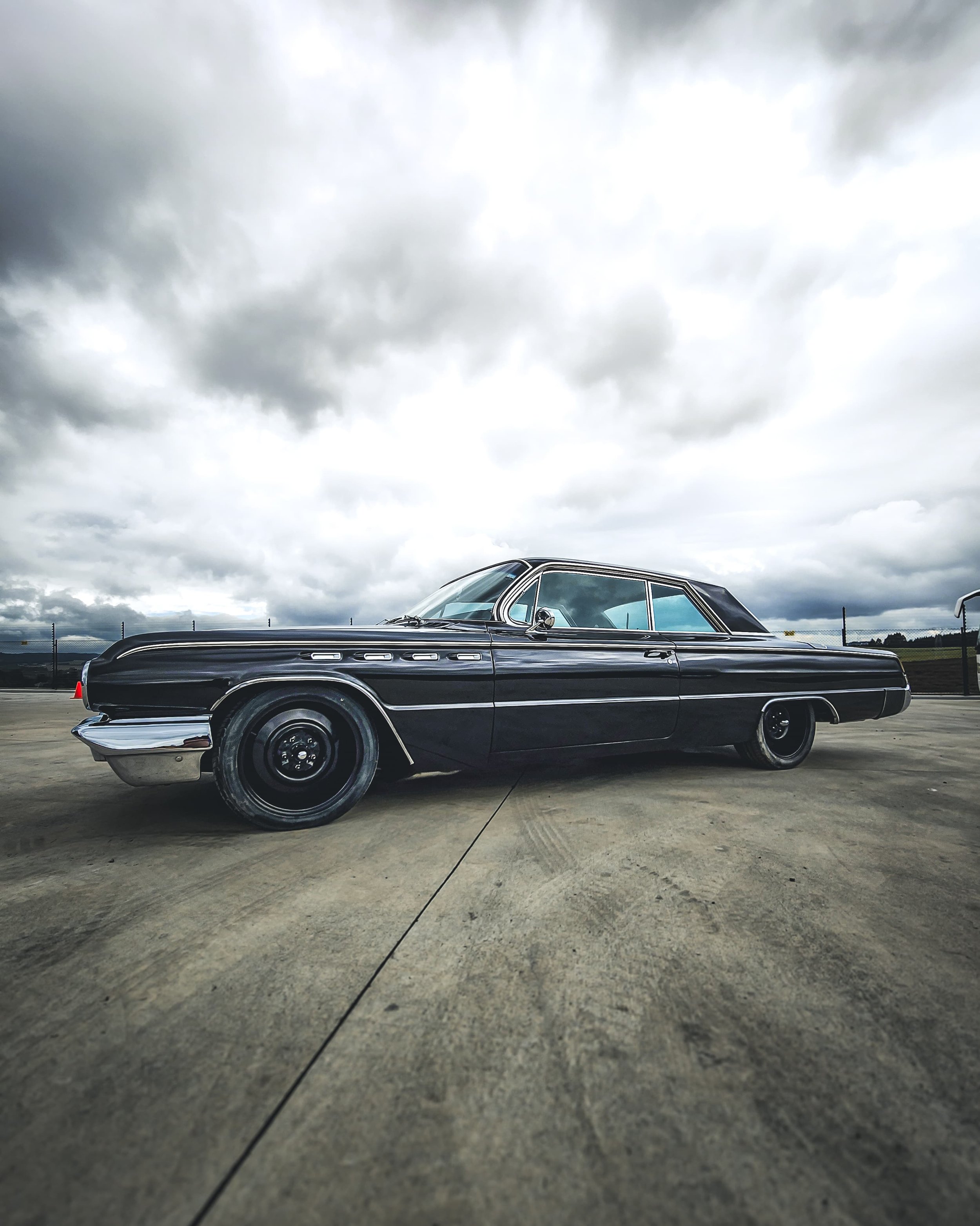 A vintage black car parked on a concrete surface under a cloudy sky.