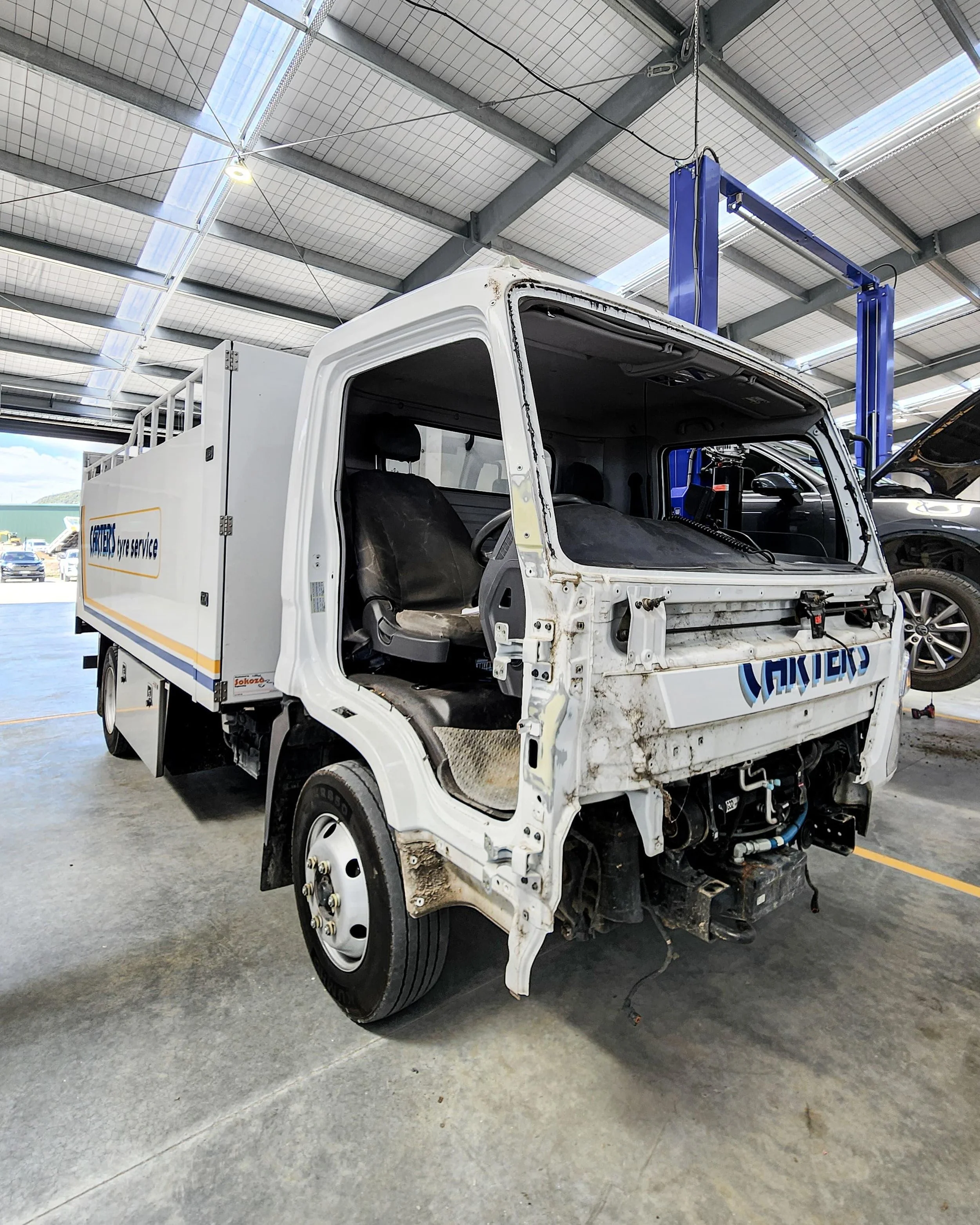 White utility truck with front panel missing, exposing the driver's seat and internal components, inside a warehouse or garage.