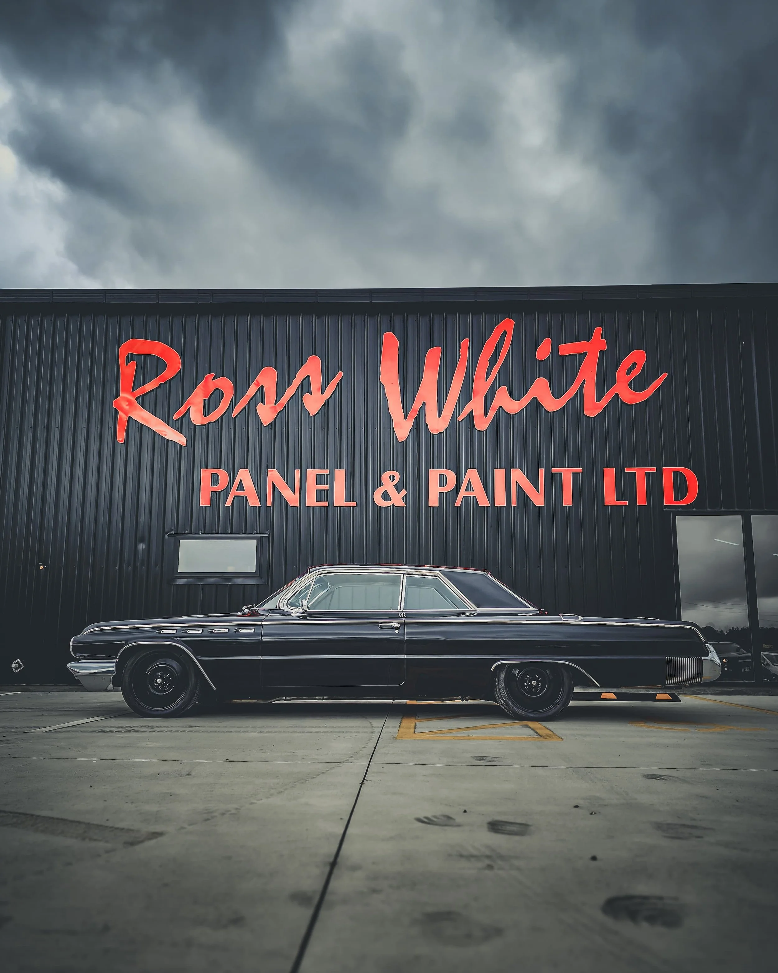 Sign reading 'Ross White Panel & Paint LTD' on a black building with a classic black car parked in front and dark stormy clouds in the sky