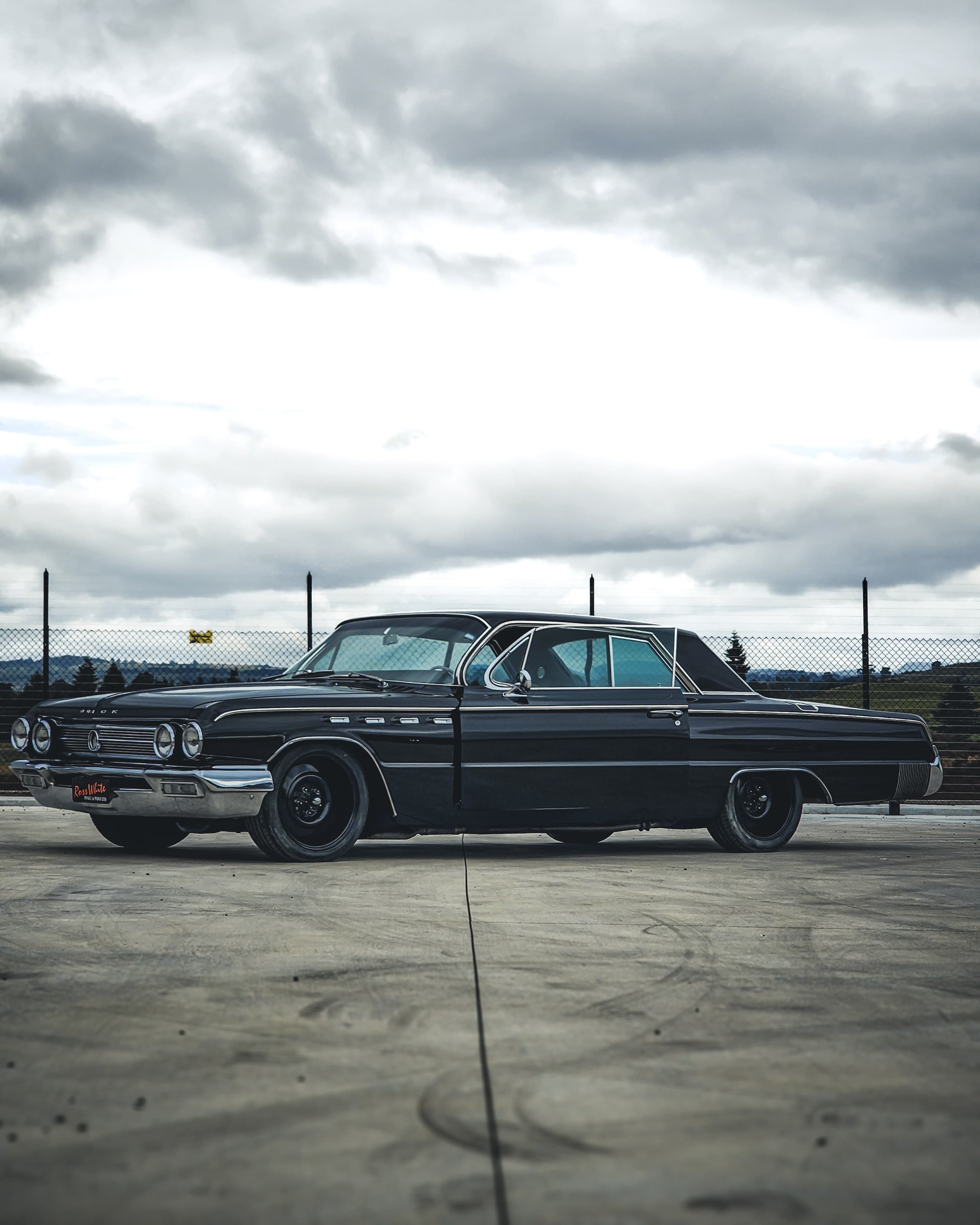 A vintage black car parked on a concrete surface with a cloudy sky and a chain-link fence in the background.