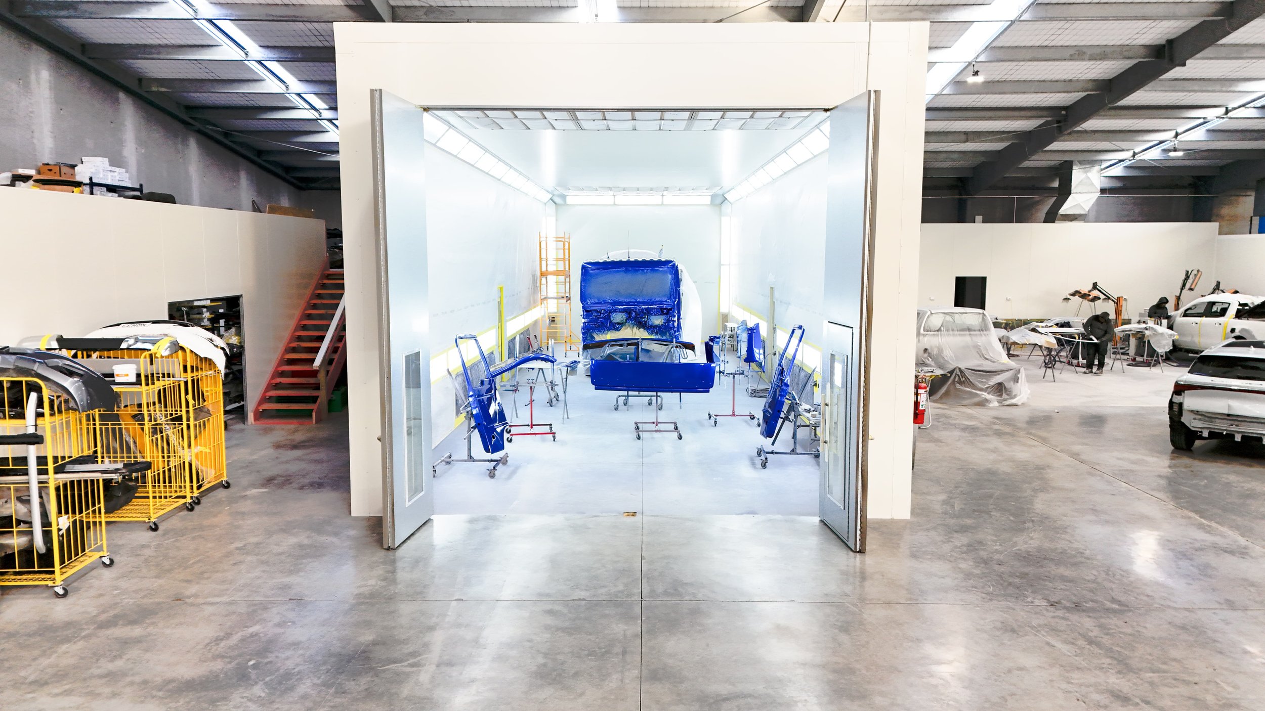 Automotive body shop with a car frame in the spray booth and various car parts and vehicles around the workspace.