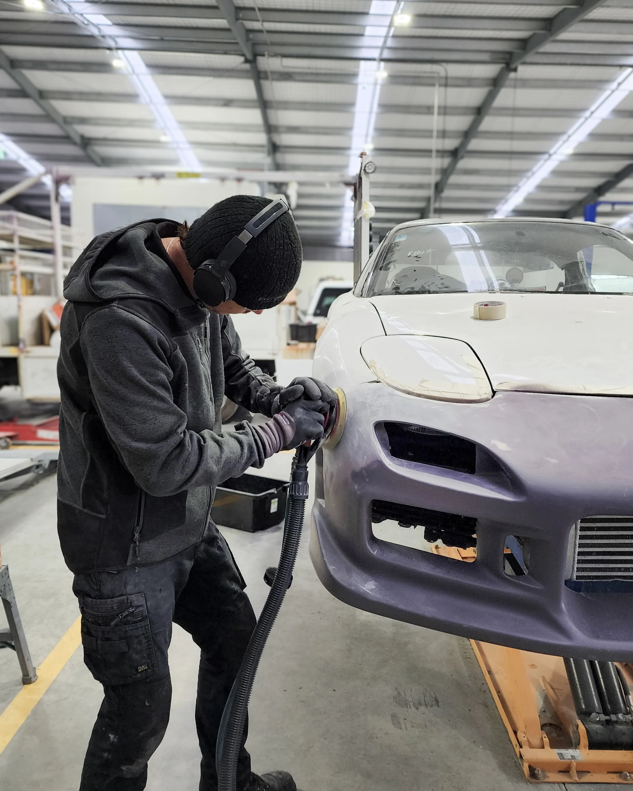 A person wearing a black beanie, headphones, black gloves, and a dark hoodie is polishing a silver sports car with a handheld polishing machine inside a workshop.