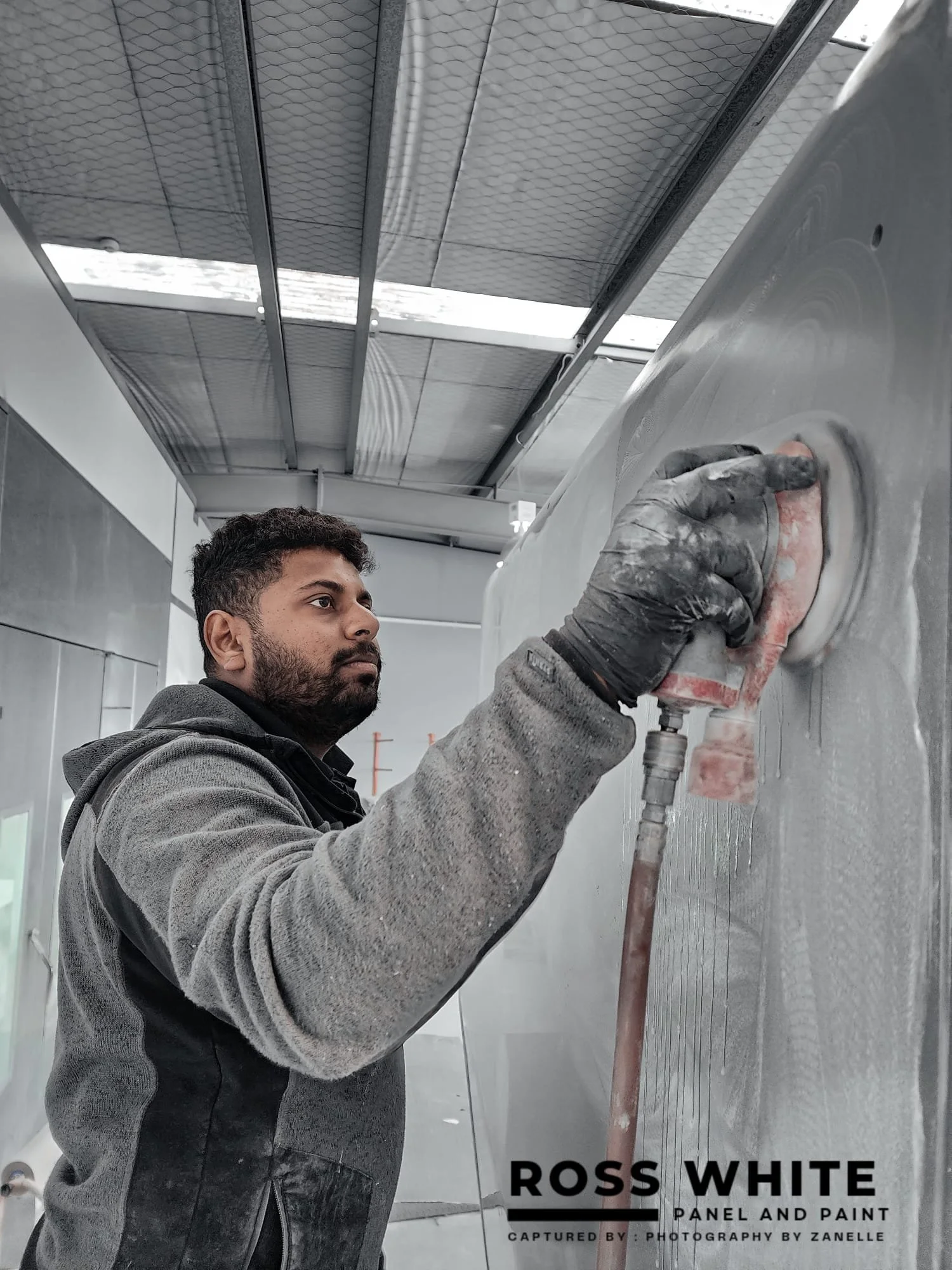 A man wearing a gray hoodie and black gloves is polishing or sanding a large metal panel with a power tool in an industrial setting.