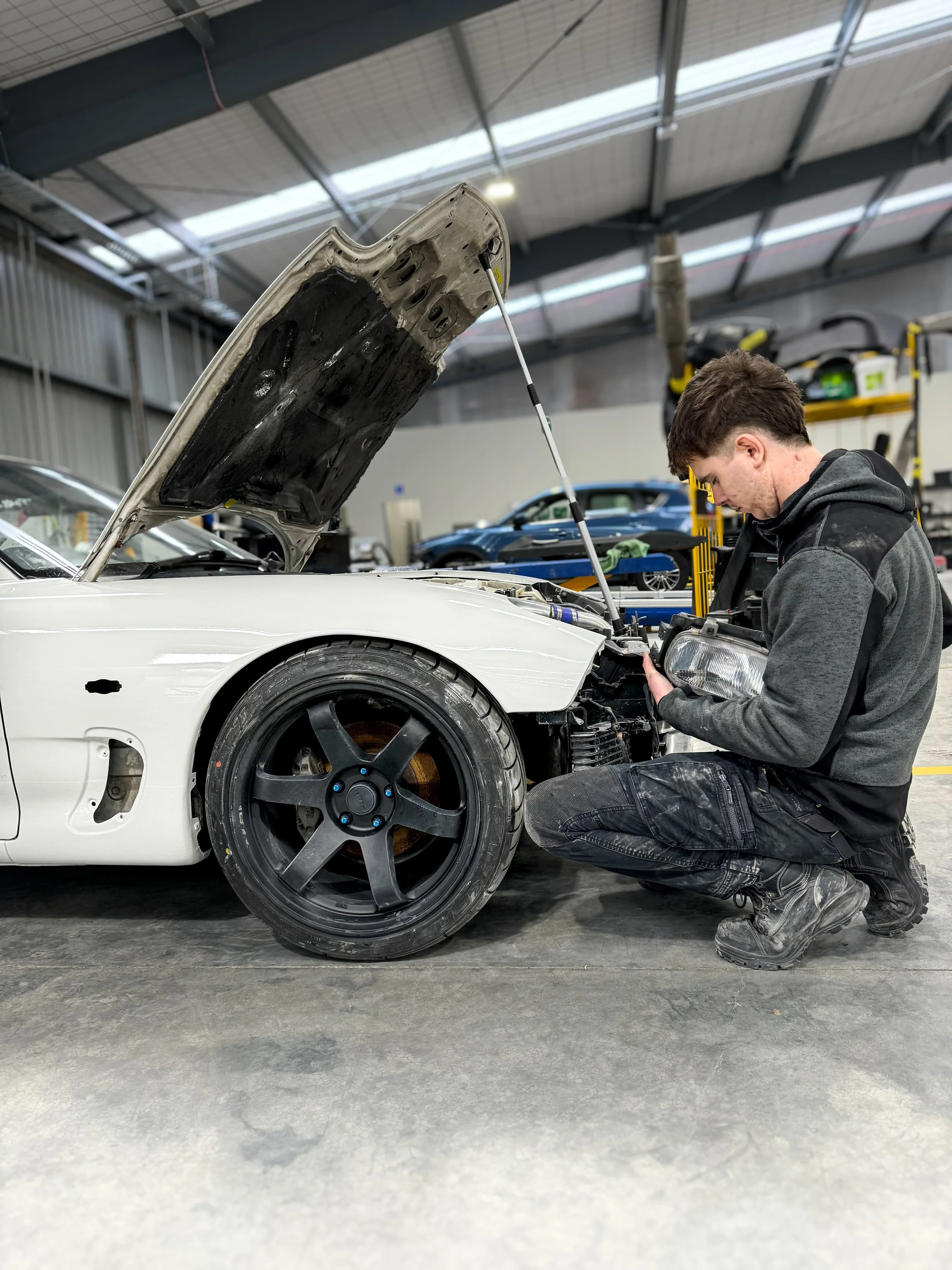 A man kneeling in front of a white car with an open hood, working on the engine inside a car repair shop.