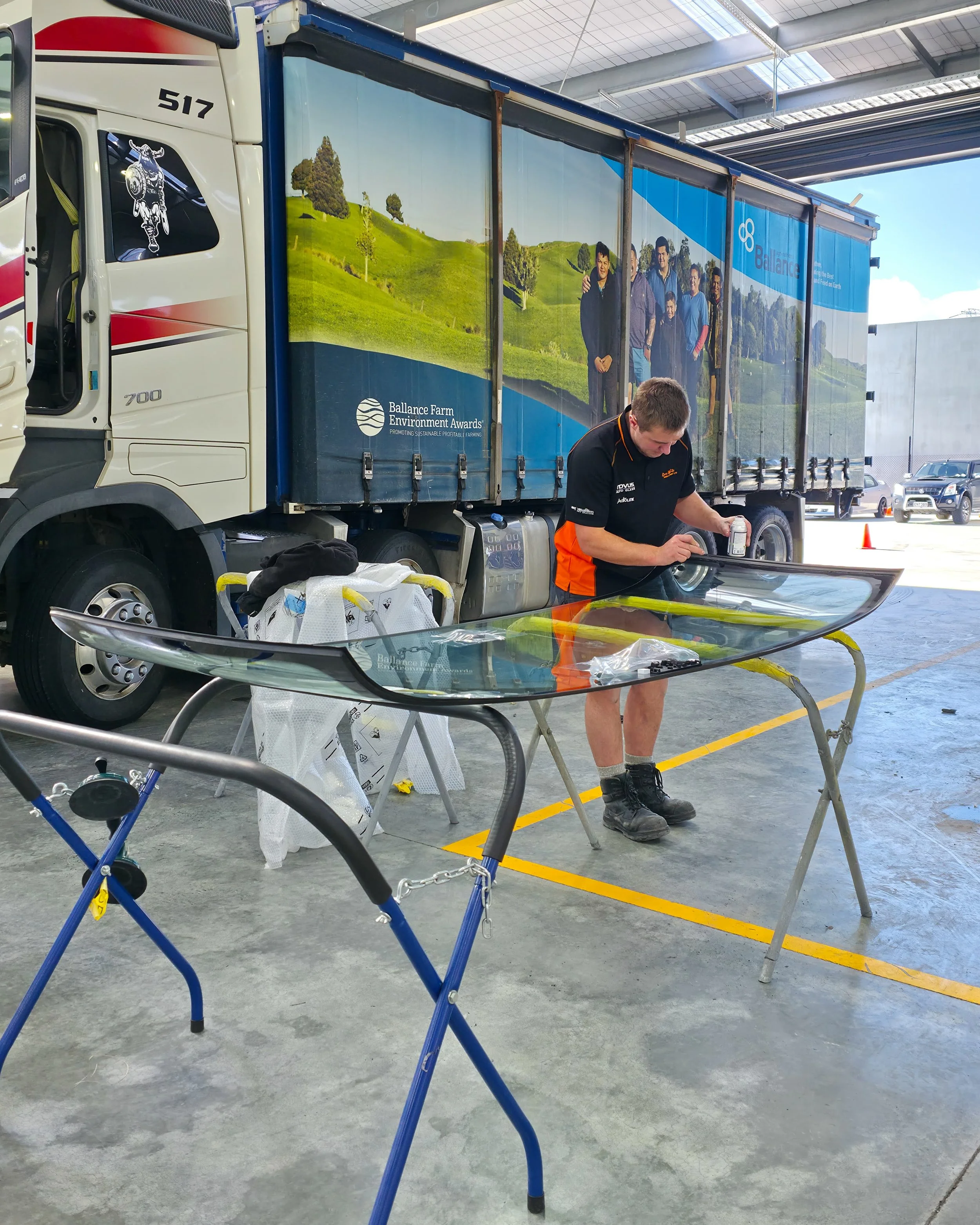 A man working on a car windshield in a warehouse or garage, with a large truck behind him. The truck has a colorful outdoor scenery mural and the logo 'Balance Farm Environment Awards'. There are various tools and a workbench nearby.