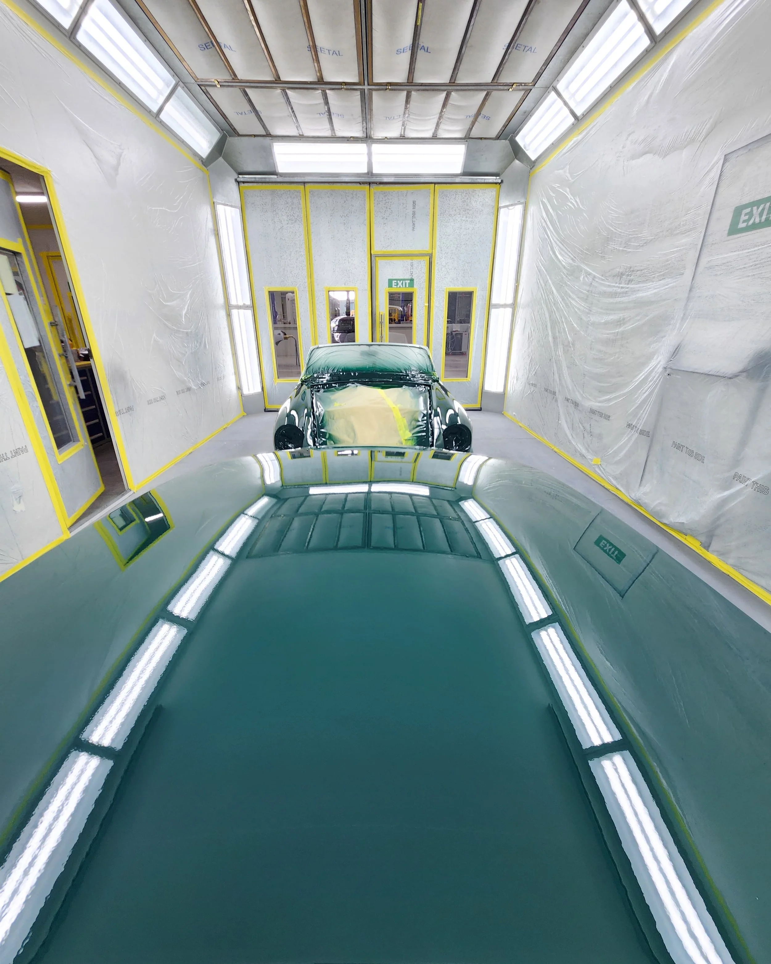Indoor car repair or painting booth with partially painted cars, bright fluorescent lighting, and plastic sheeting on the walls.