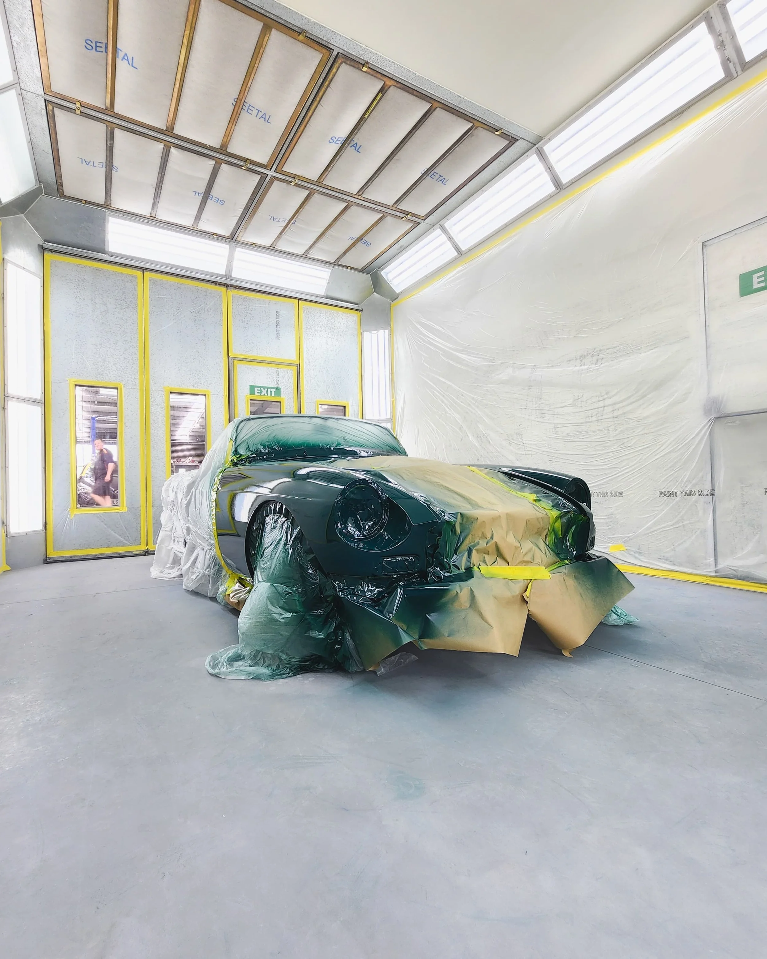 A black sports car in a paint booth, partially covered with masking paper and plastic for painting or detailing.