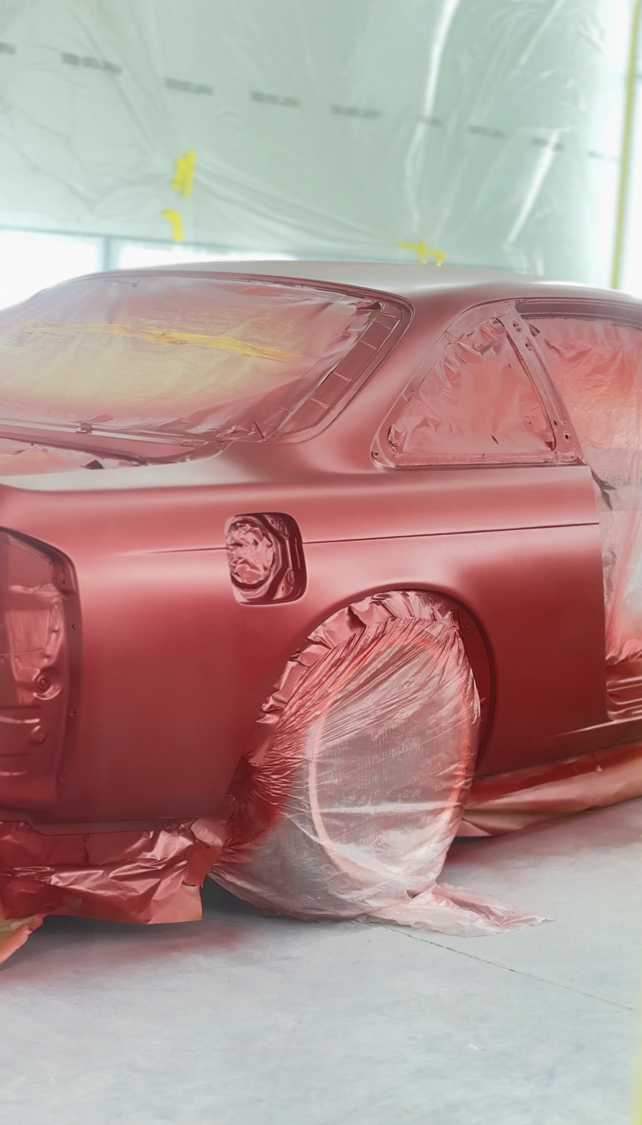 A car is being painted with a pinkish hue inside a spray booth, with protective plastic covering the windows, wheels, and other parts.