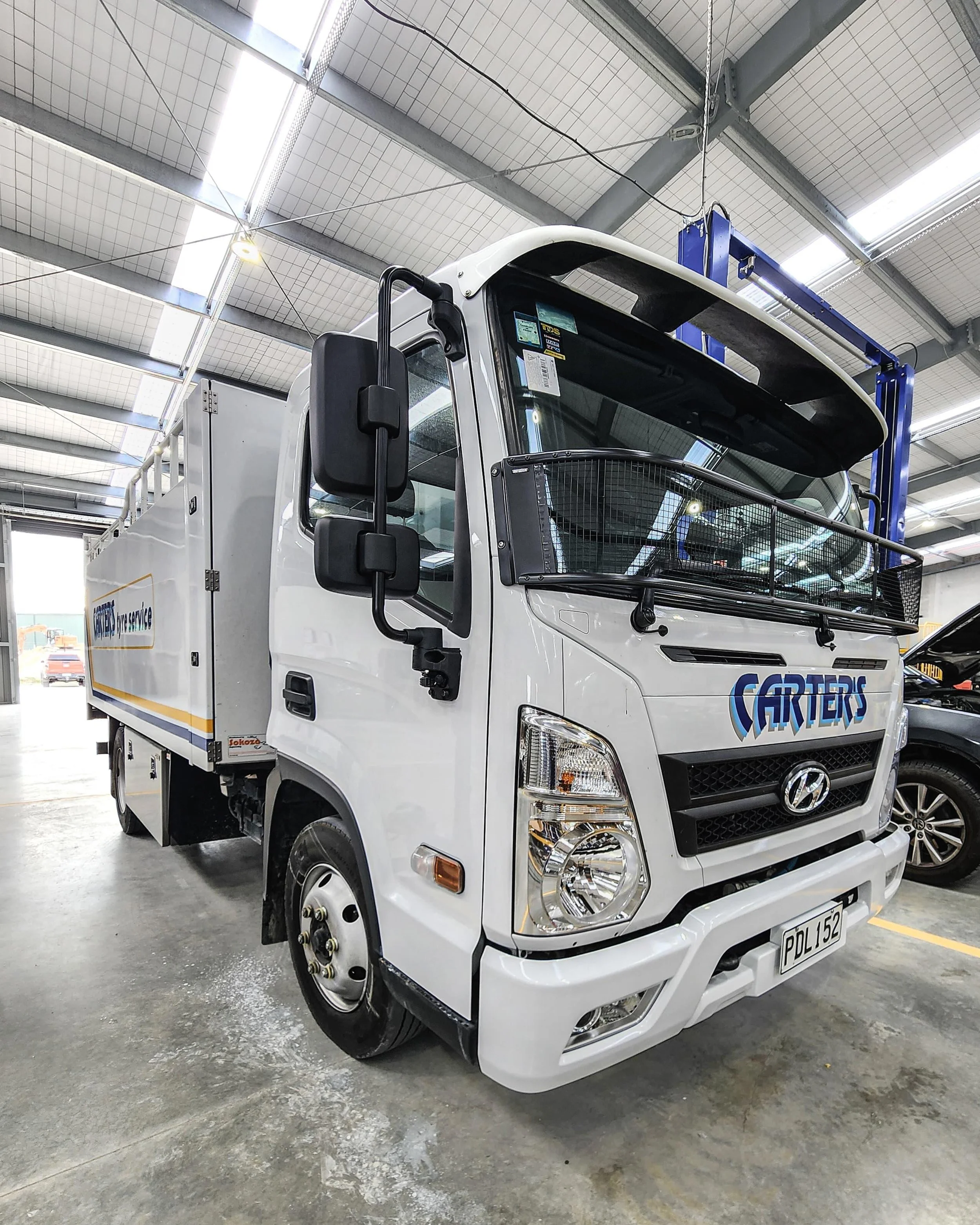 White Hyundai service truck parked indoors with a front grille, headlights, and a flatbed, bearing a 'Cartels' logo on the front and side, inside a garage or warehouse.