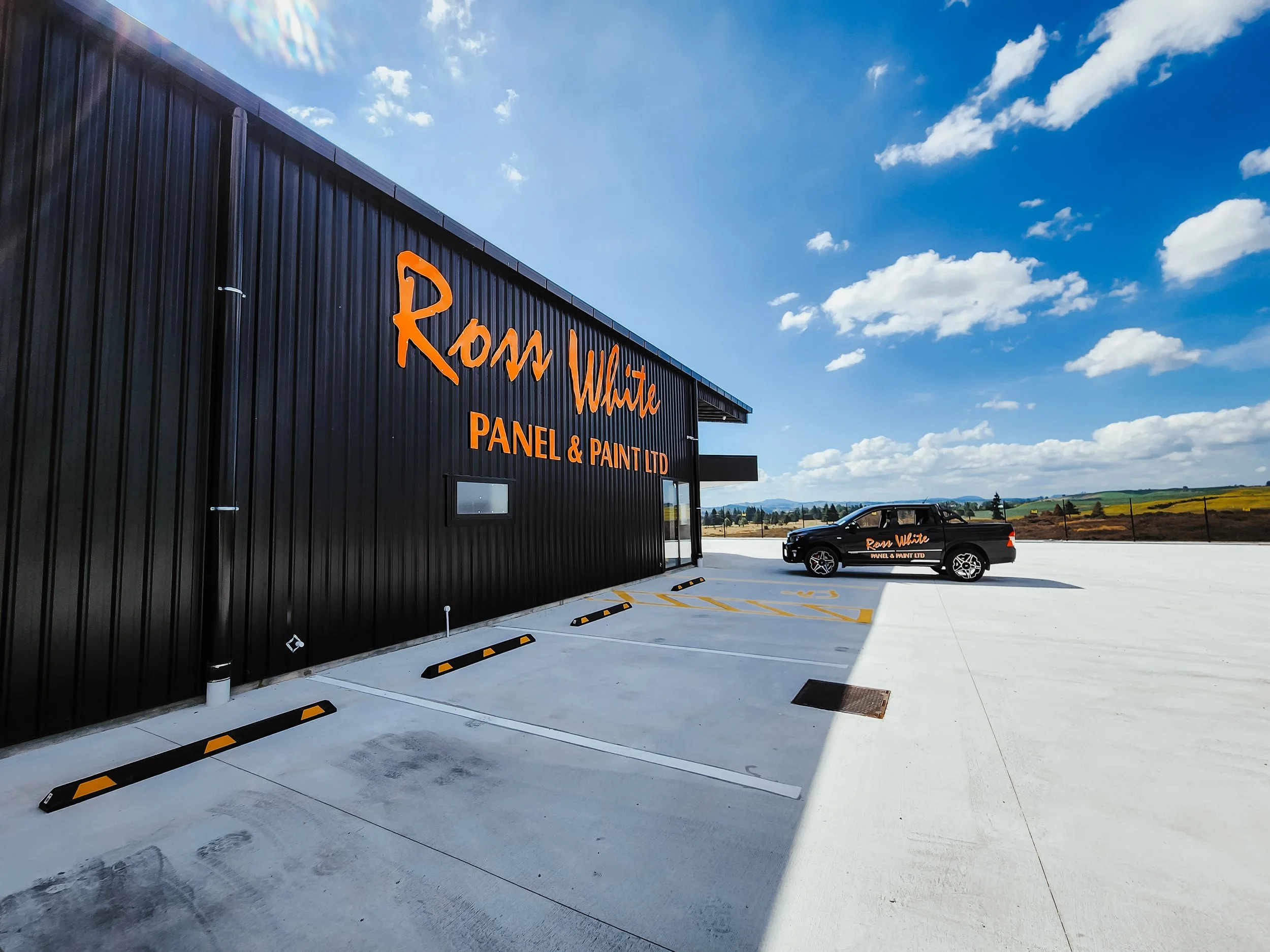 A black building with orange lettering that reads 'Rom White Panel & Paint Ltd,' and a black pickup truck parked in front of it on a large white concrete lot. The sky is partly cloudy with a scenic landscape in the distance.