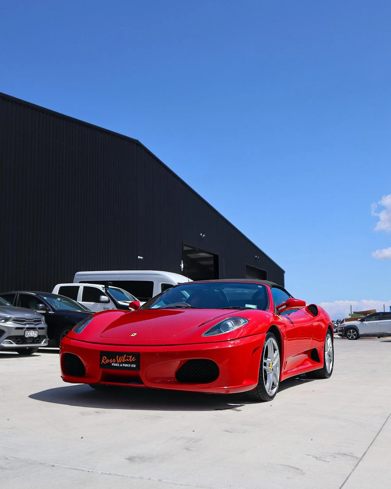 Red Ferrari sports car parked in an outdoor lot with other vehicles and a black industrial building in the background.