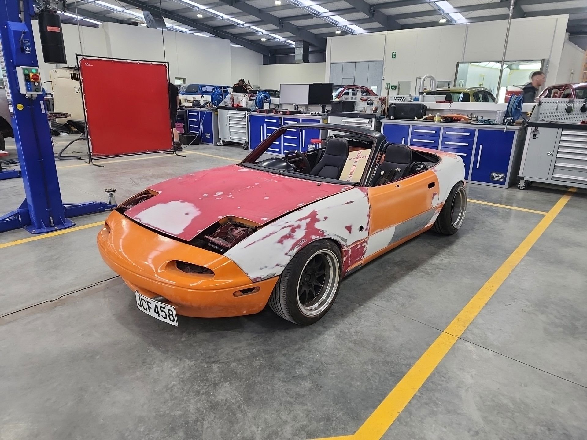 An unfinished orange and white convertible sports car in a garage workshop, with the paint partially applied and masking tape still visible on the body. Behind the car, there are blue tool cabinets, workbenches, and various automotive equipment.