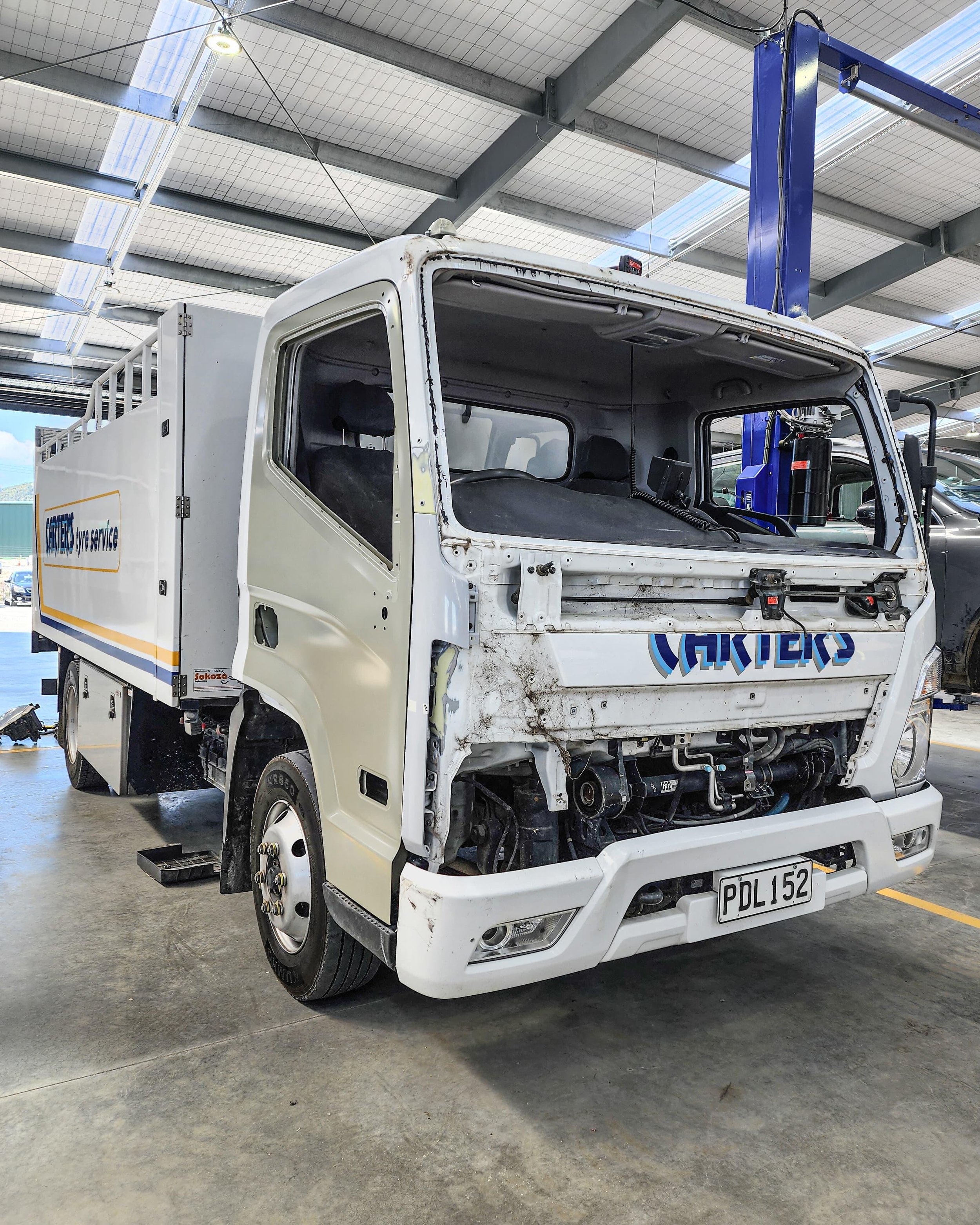 A white delivery truck with the front panel removed, showing exposed engine components and damage, parked inside a warehouse or workshop.