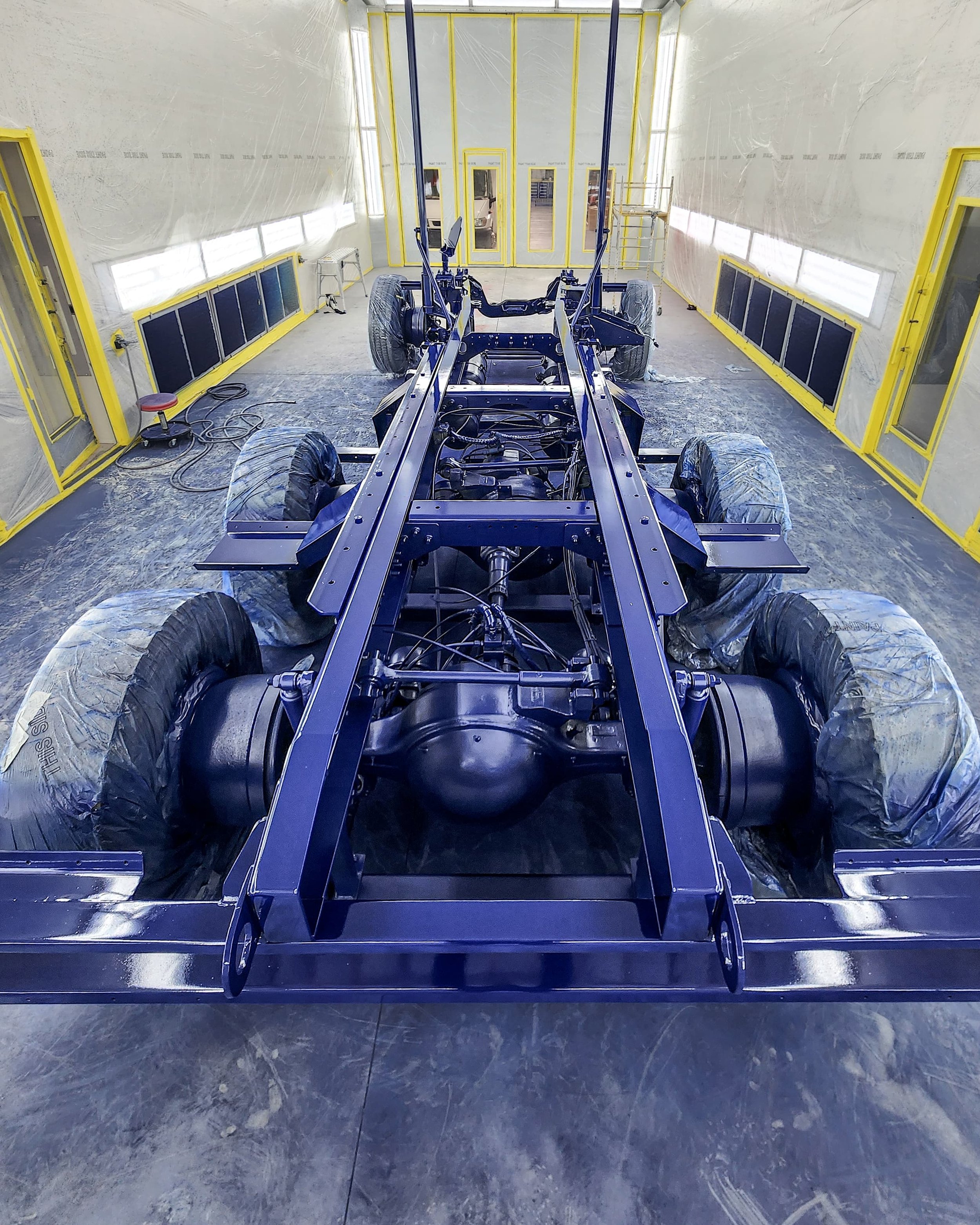 View of a vehicle chassis inside a paint booth, with wheels and suspension components visible, and yellow framing around the openings of the booth.
