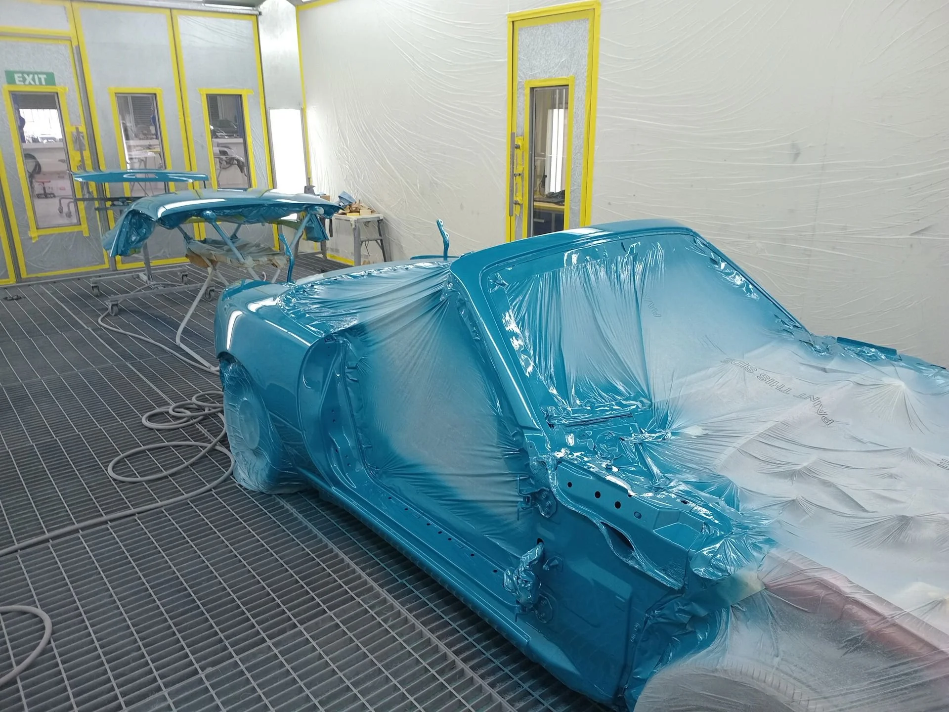 A car in a paint booth, masked and covered with plastic for painting, with a small parts frame in the background also masked for painting.