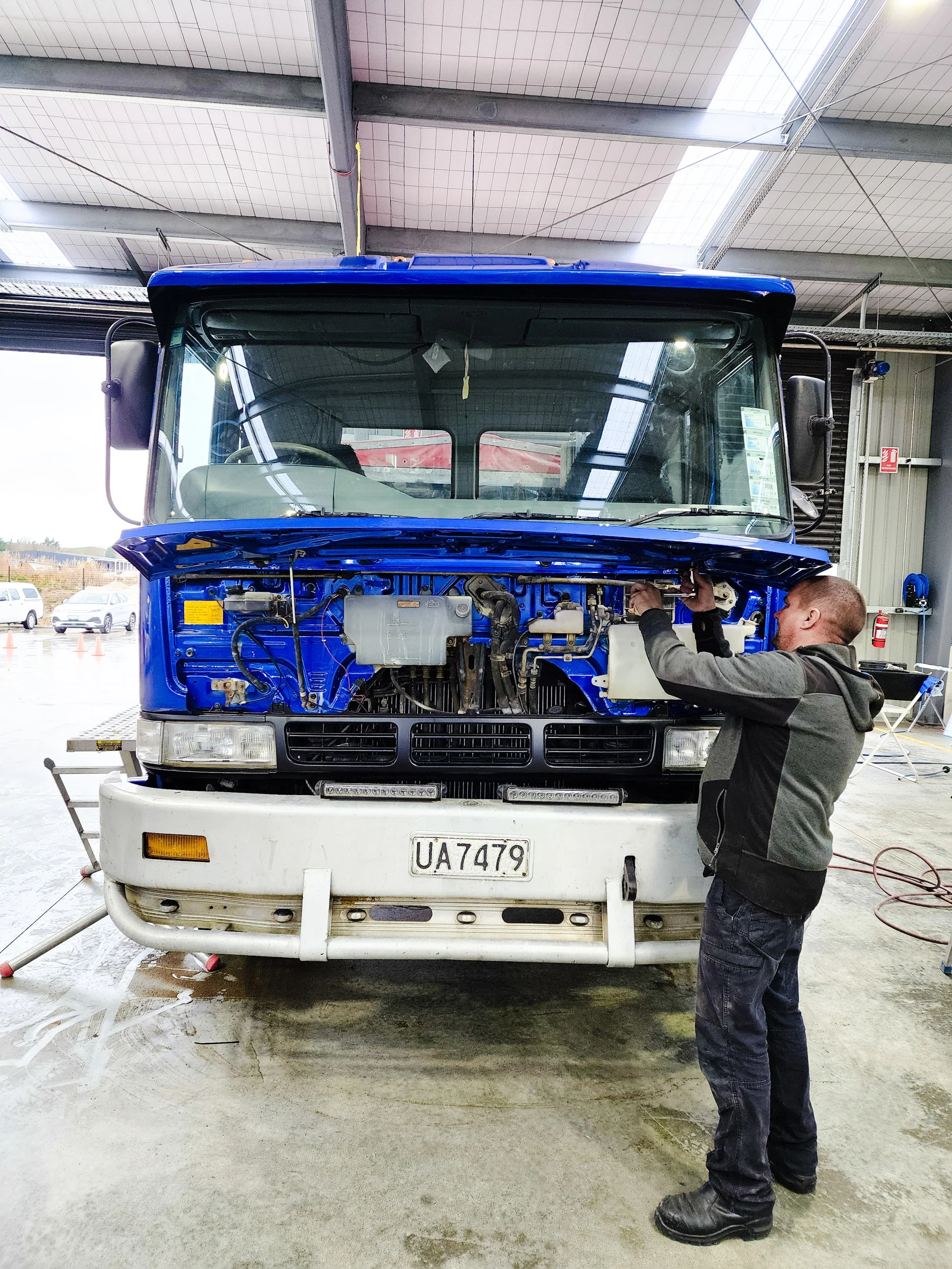 A mechanic working on a blue and white truck inside a garage, with the hood open and the engine exposed.