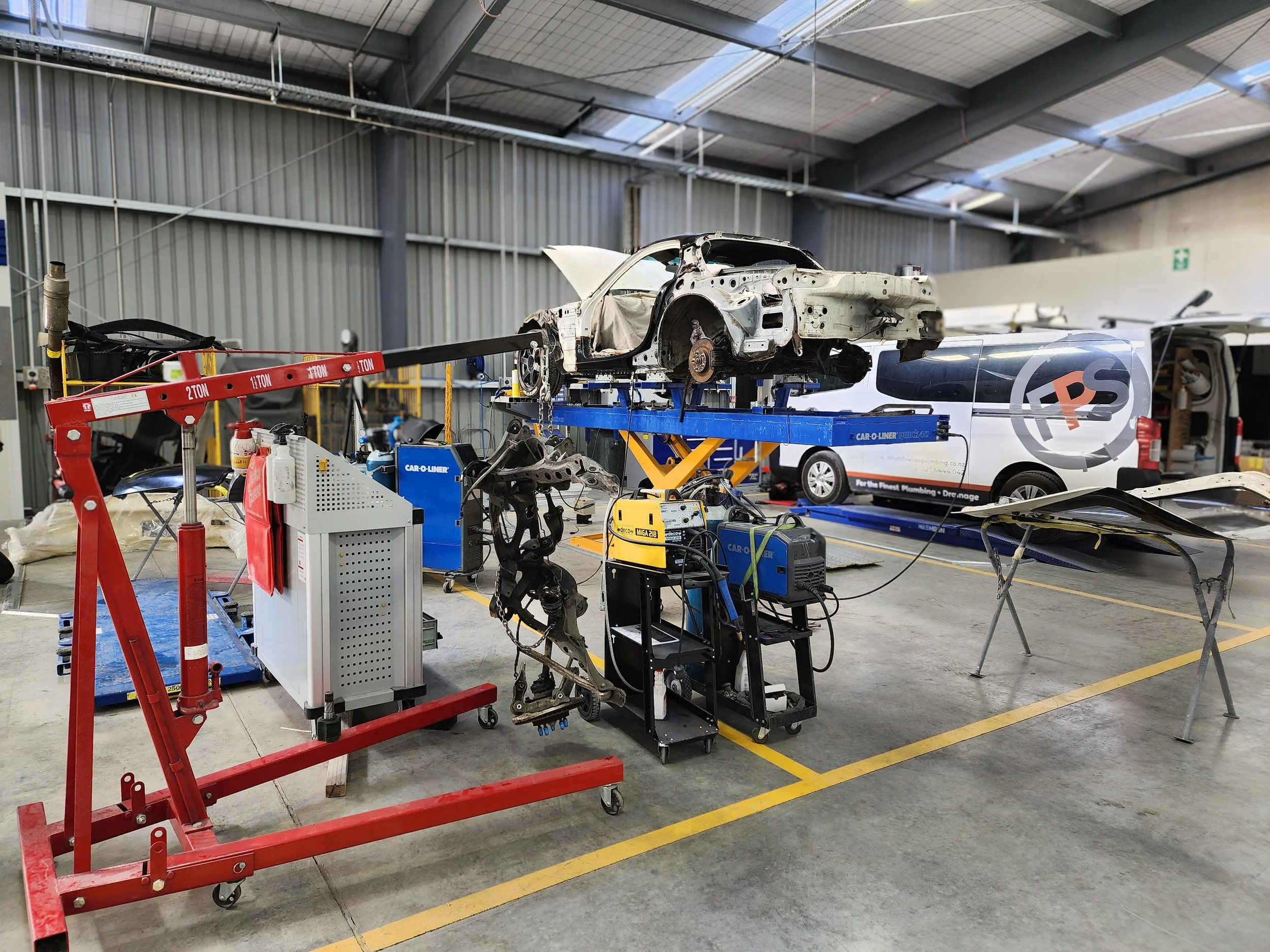 Automotive workshop with a damaged car on a lift, various repair tools, and equipment, with another vehicle in the background.