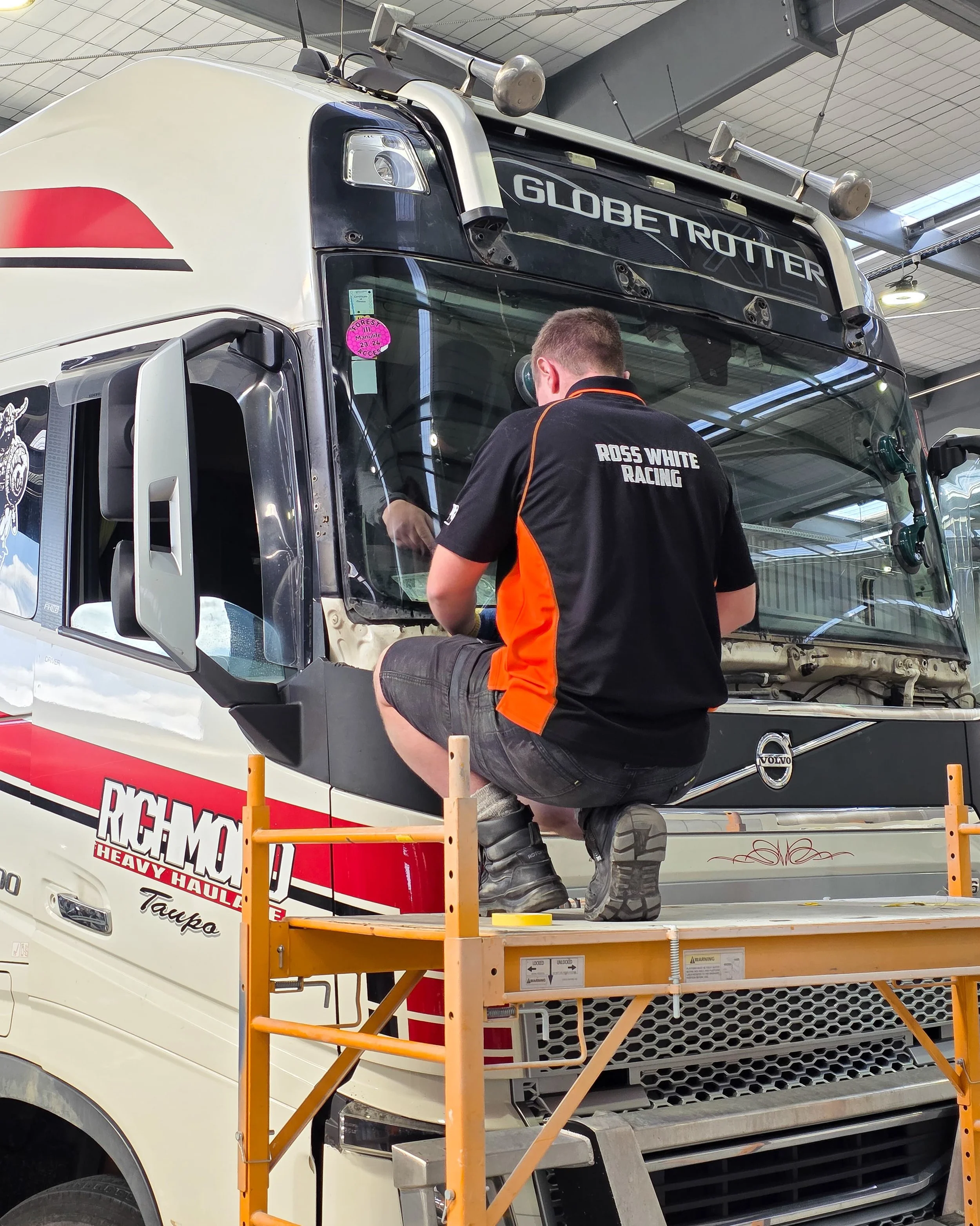 Person in Racing team uniform working on a large semi-truck at an indoor facility.