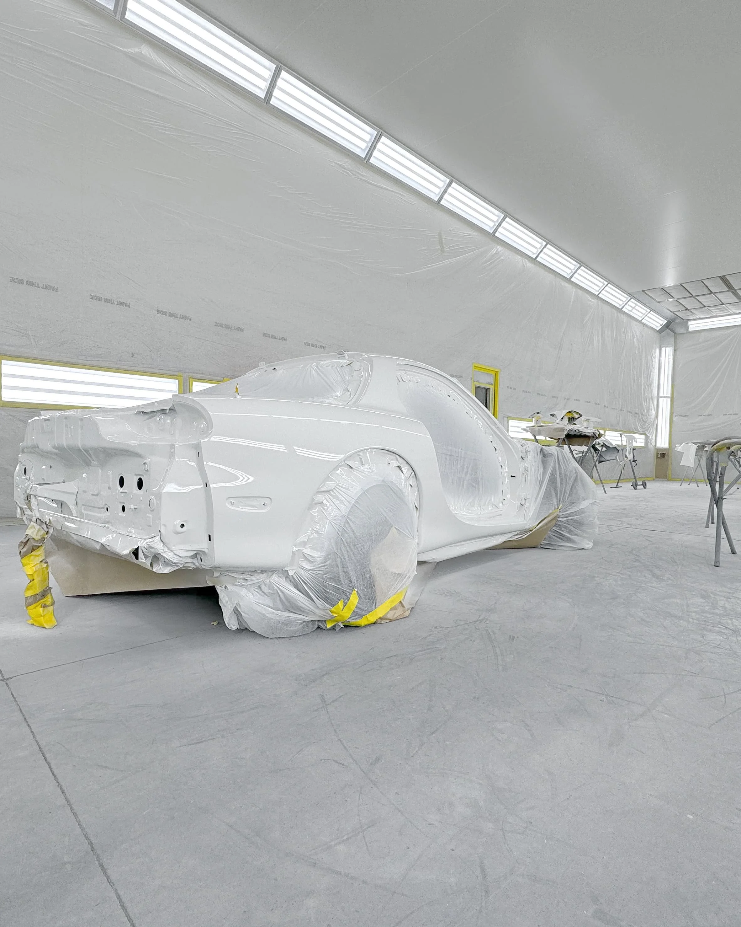 A white sports car body on a stand in a well-lit auto body shop, mask and paper coverings over wheels and parts, with work tables and bright fluorescent lighting.