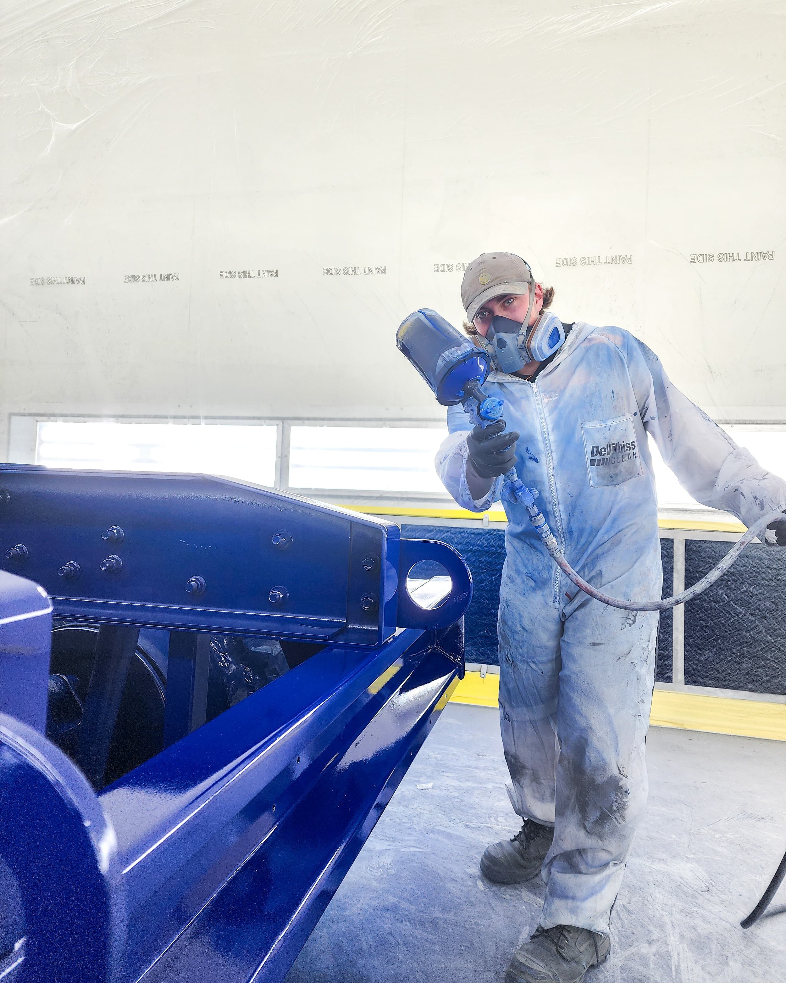 Worker with a respirator mask spray-painting a blue metal structure inside a workshop.