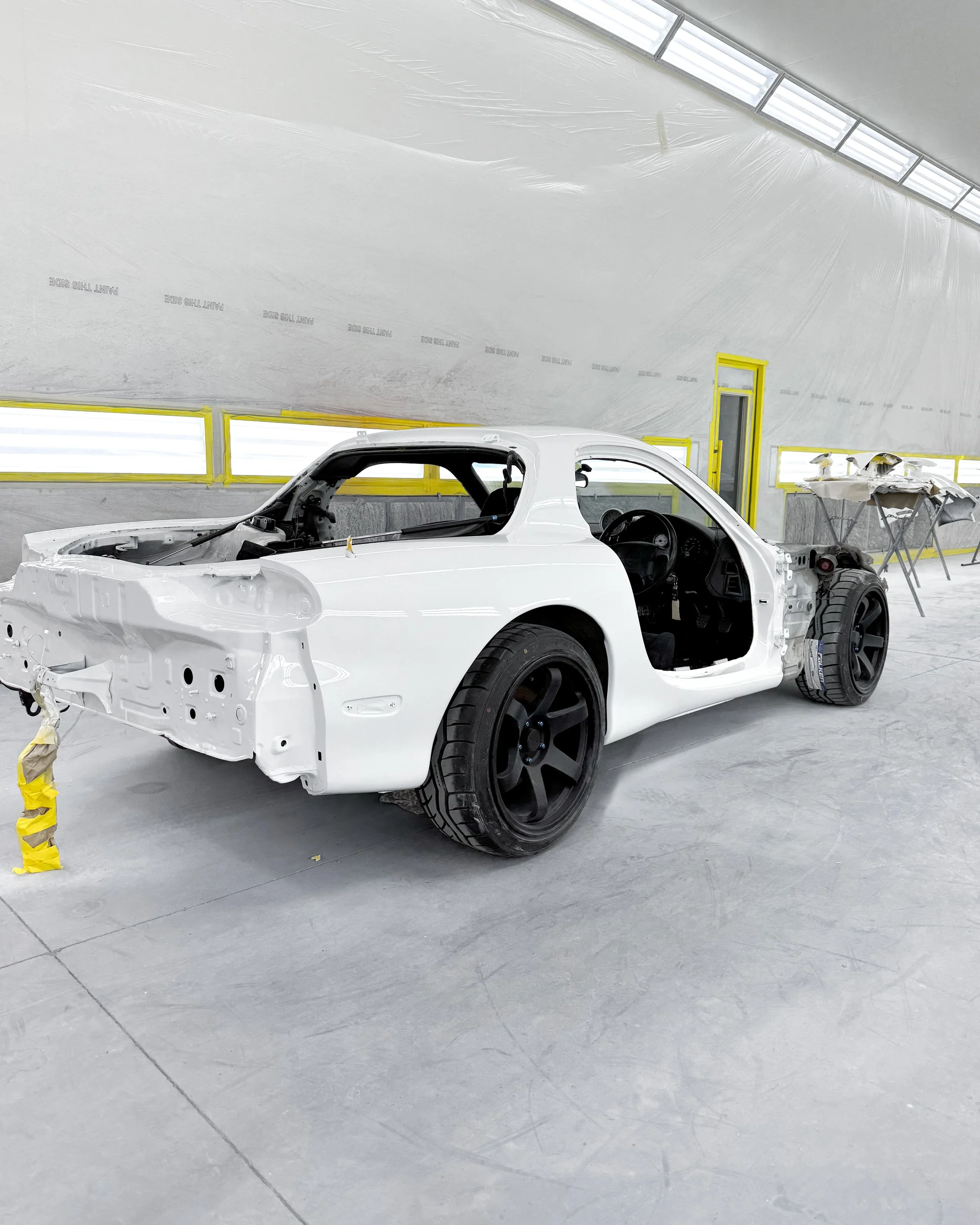 A partially assembled white race car with black wheels in an industrial workshop with yellow safety markings and tools in the background.