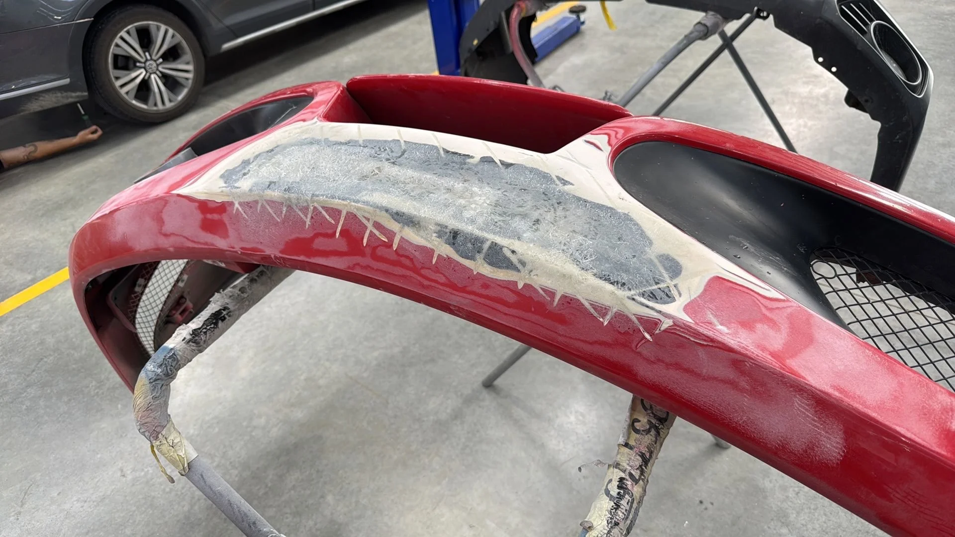 Close-up of a damaged red vehicle body or part, possibly from a race car, with visible scratches, scuffs, and taped areas for repair, placed on a stand in a workshop or garage.