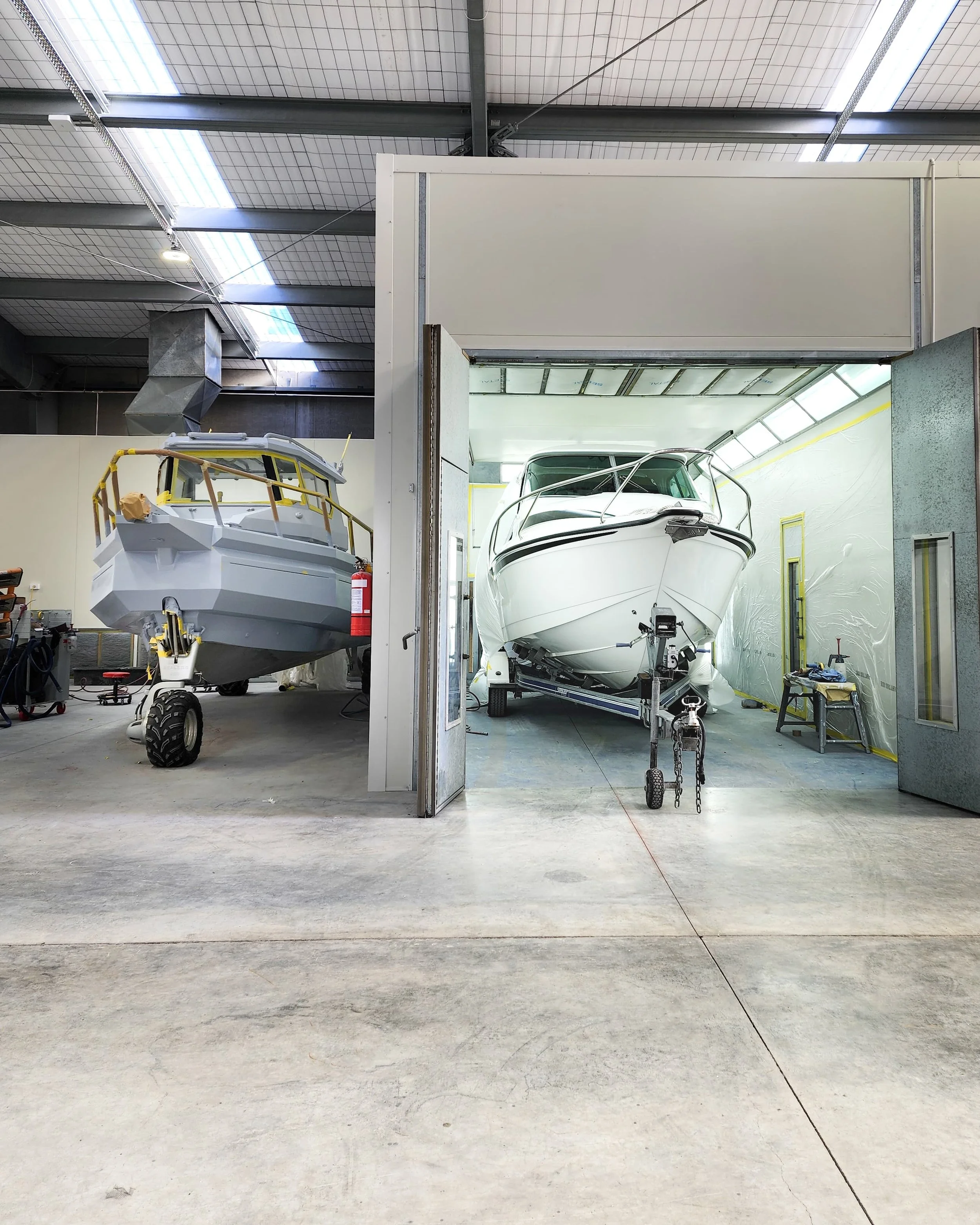 Two boats are stored in a boat maintenance or assembly shop. One boat on the left is painted gray and on wheels, the other on the right is white and on a trailer.