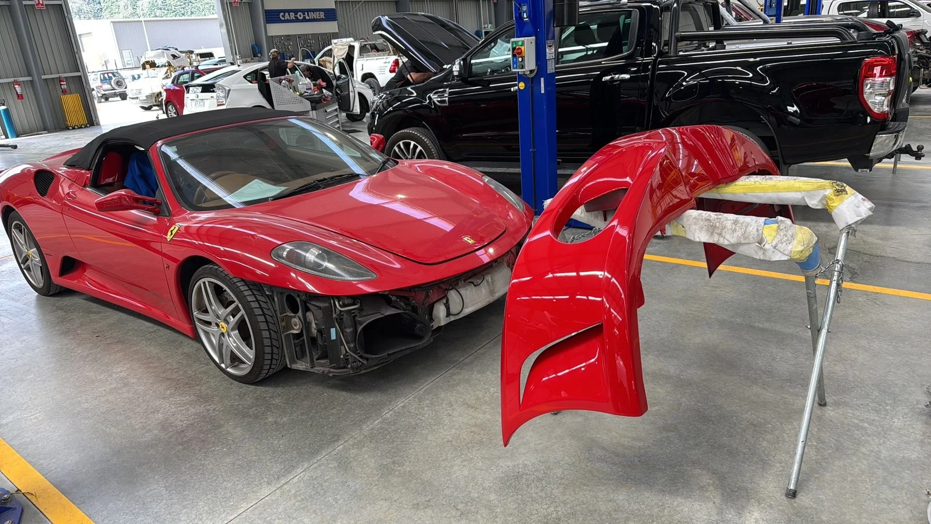 A red Ferrari sports car is undergoing repairs in an auto body workshop. The front bumper is removed and placed on a stand nearby, while the hood and other parts are being worked on. Other vehicles are visible in the background.
