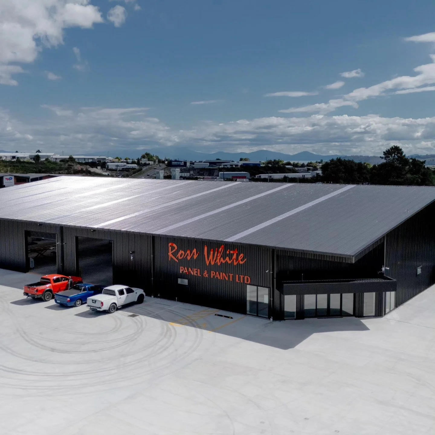 A large industrial building with a metallic roof and black walls, with a sign that reads "Ron White Panel & Paint LTD." Three trucks—red, blue, and white—are parked outside, with tire marks visible in the light-colored parking lot. In the background,