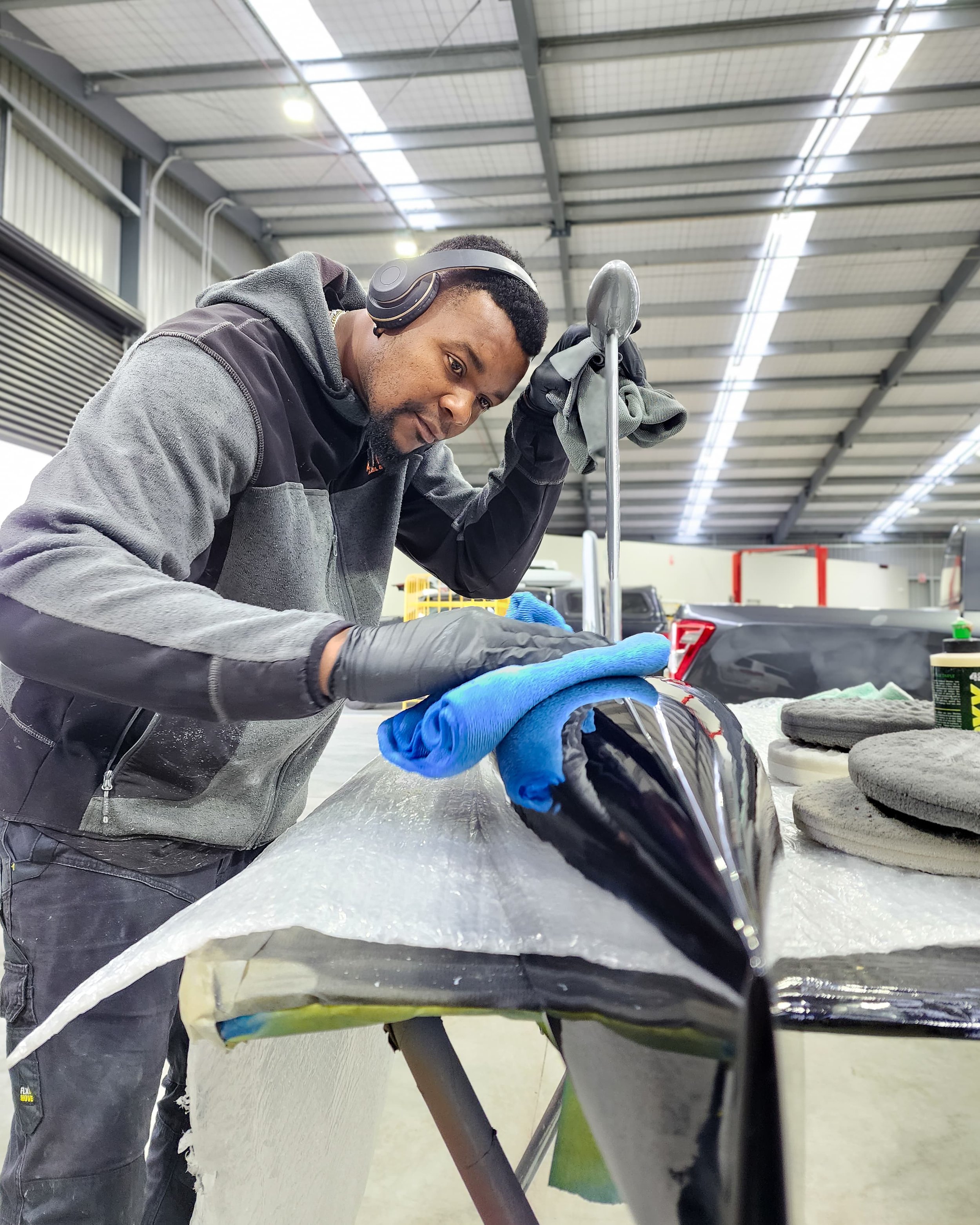 Man polishing a black motorcycle tank in a workshop, wearing gloves and headphones