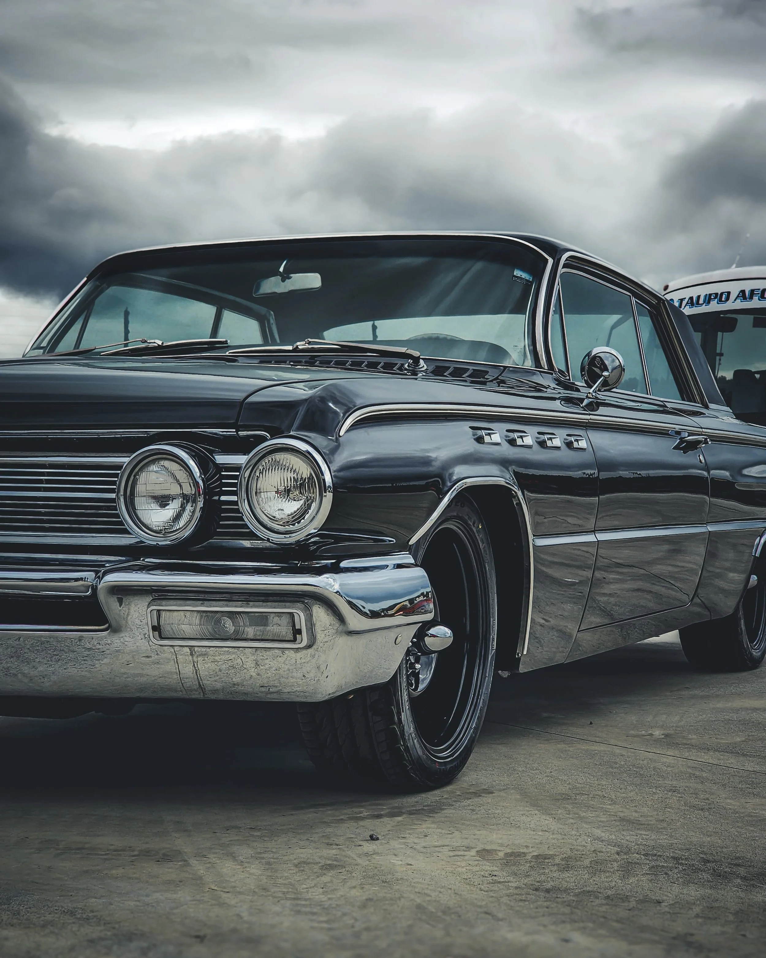 A vintage black car with chrome detailing and dual headlights parked on a concrete surface under cloudy skies.