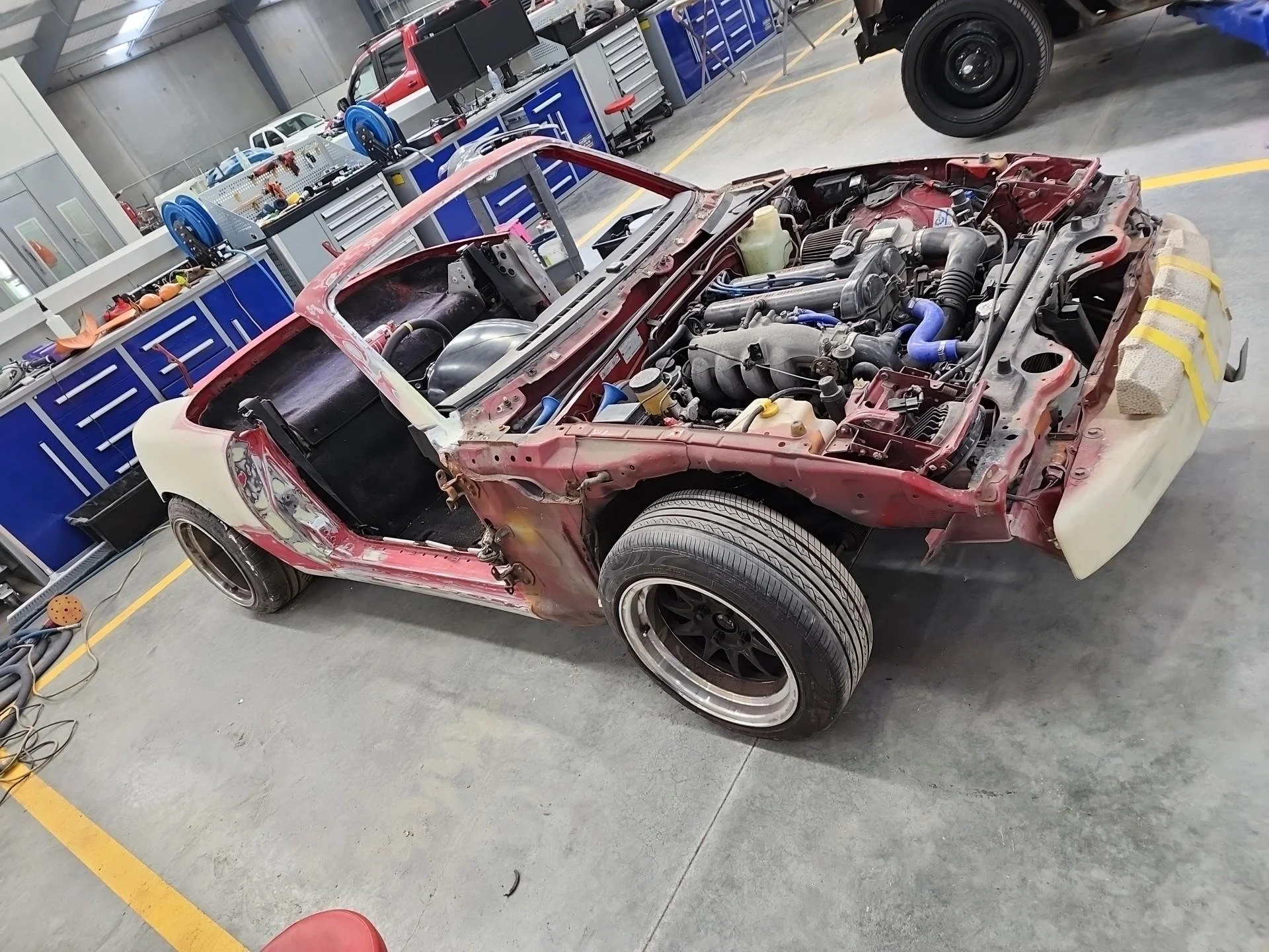 A partially disassembled car in a workshop, with its body separated from the engine bay and various tools and equipment around.