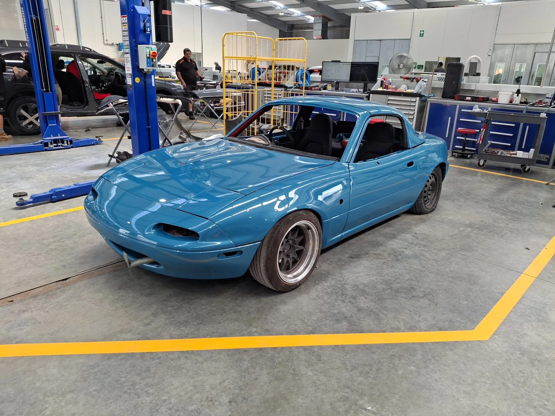 A partially assembled blue Mazda MX-5 Miata in a car workshop, with its front bumper and headlights missing, inside a repair or assembly area with tools and equipment around.