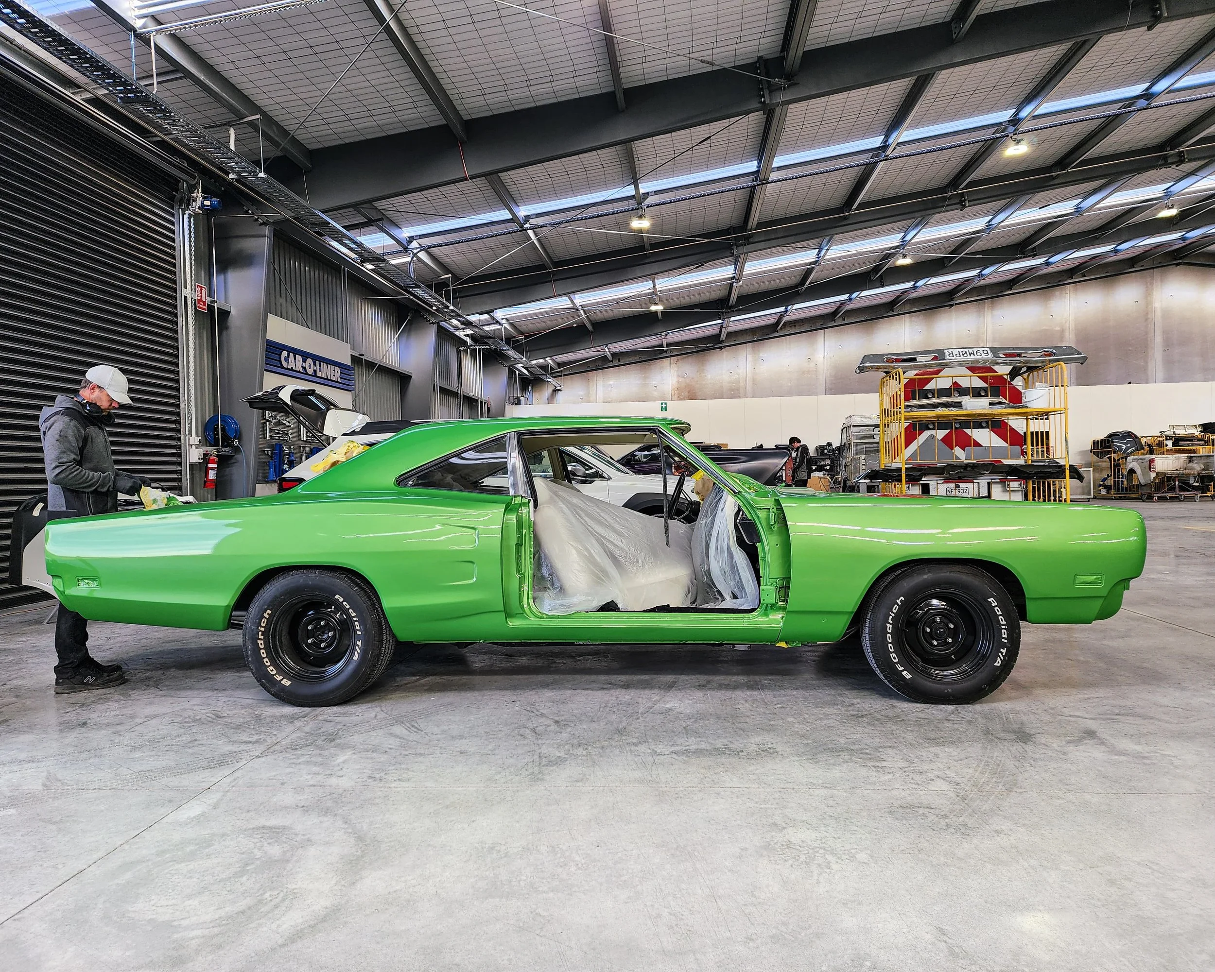 A green classic car undergoing restoration inside a warehouse, with a person working on it.